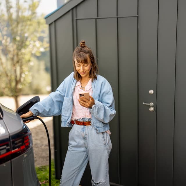 Frau lädt Elektroauto und blickt dabei auf ihr Smartphone
