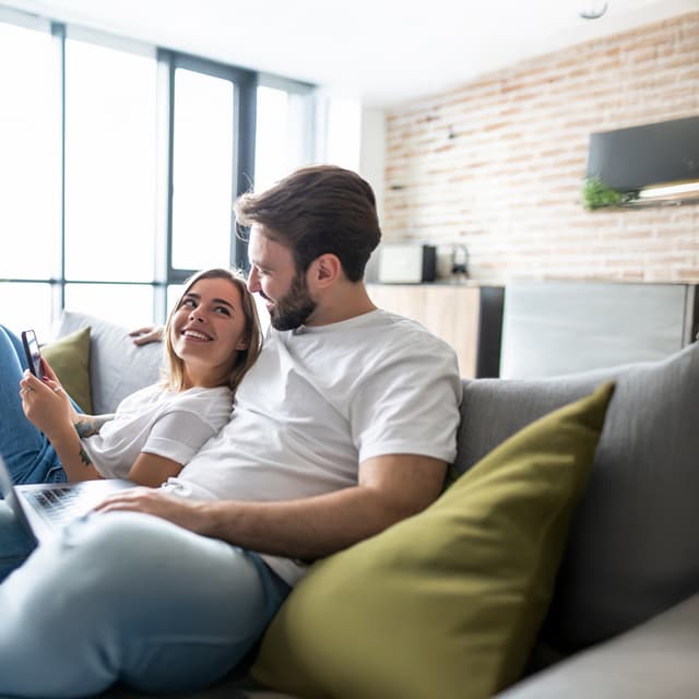 Paar sitzt auf einer Couch und lächelt sich an. Sie hält ein Smartphone in der Hand, er hat einen Laptop auf dem Schoß.