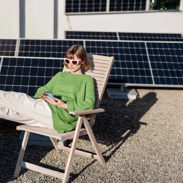 Frau sitzt in einem Gartenstuhl mit einem Smartphone in der Hand, im Hintergrund sieht man Solarpaneele