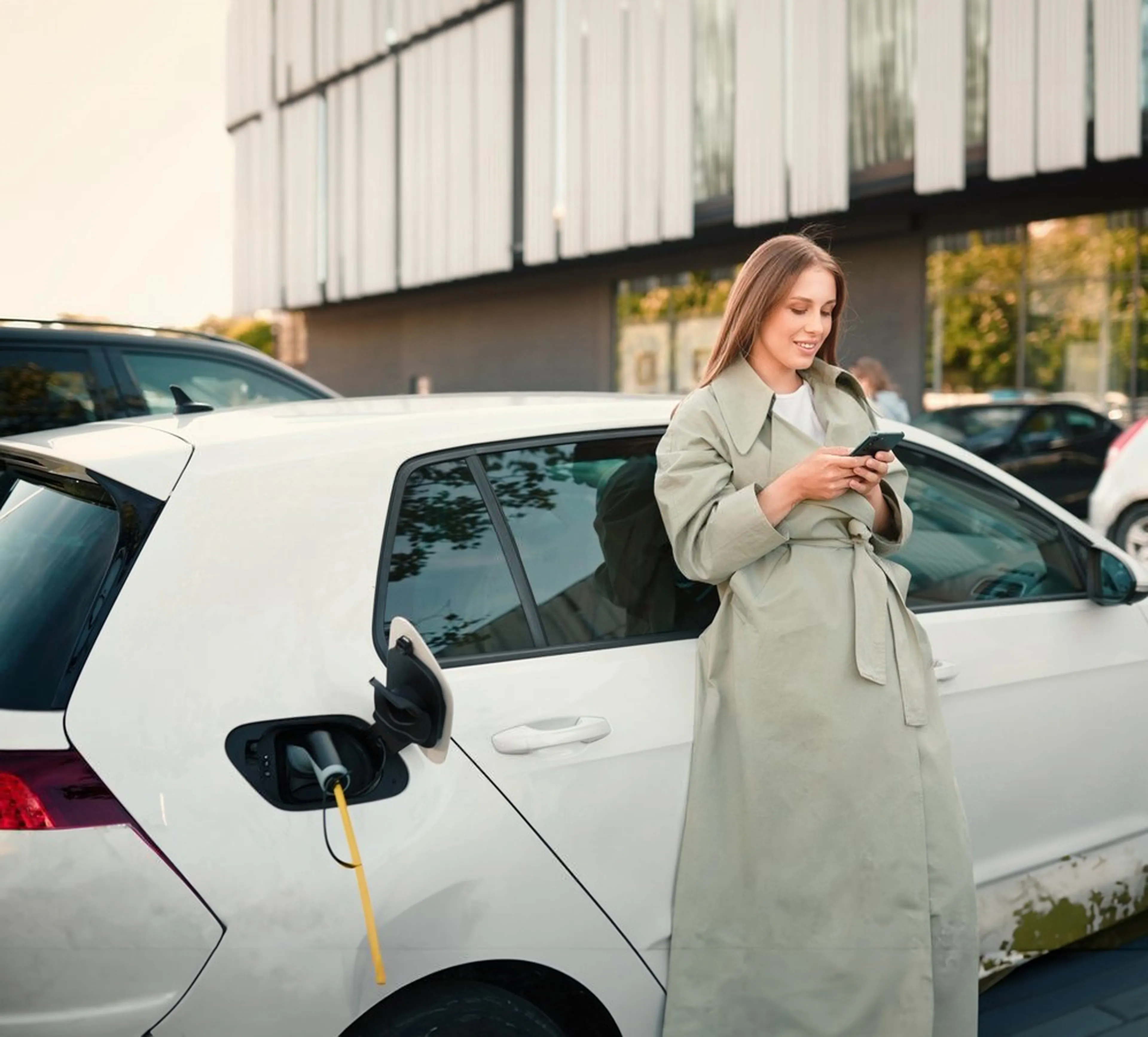 Eine Frau im Trenchcoat lehnt sich an ein weißes Elektroauto, während sie ihr Telefon benutzt. Das Auto wird an einer Station im Freien aufgeladen.
