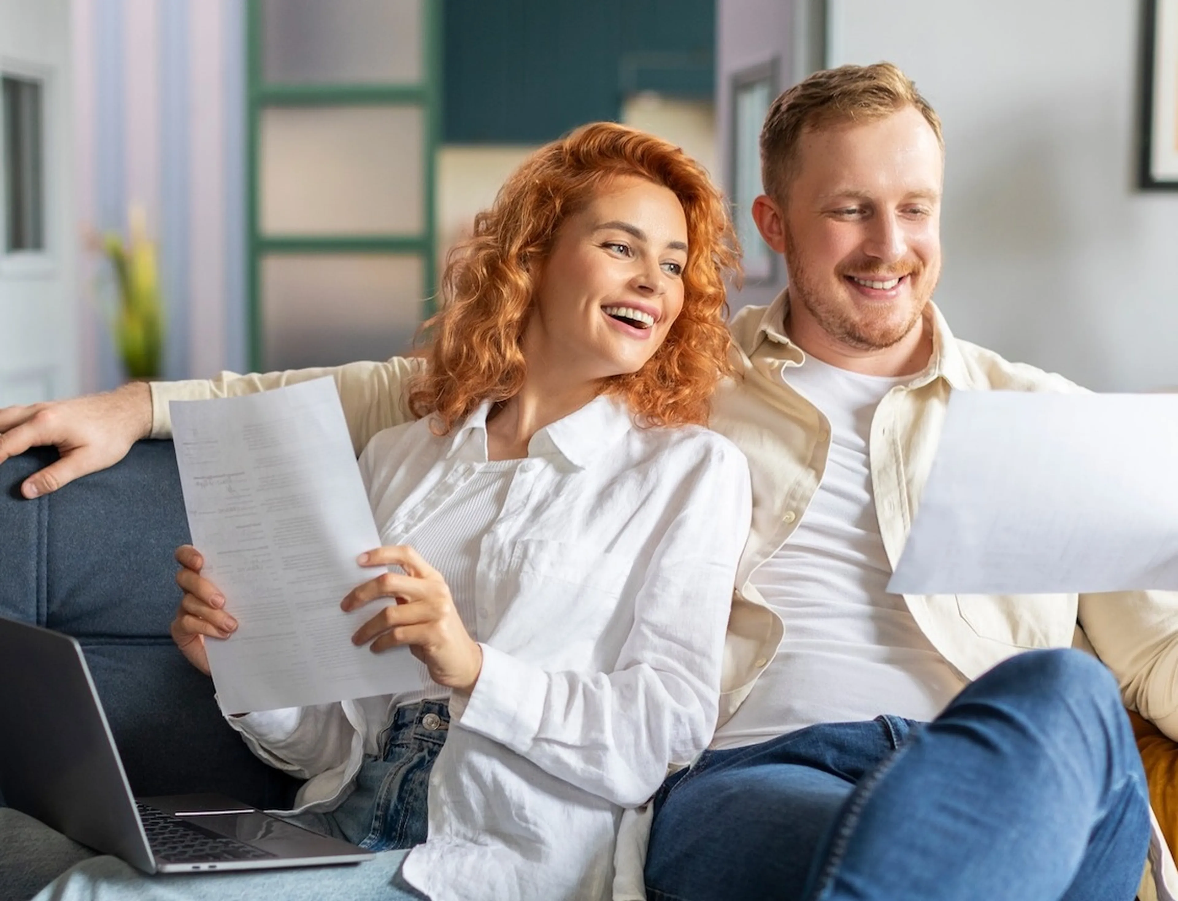 Ein Paar sitz auf dem Sofa, hält in den Händen jeweils ein Dokument und lacht.