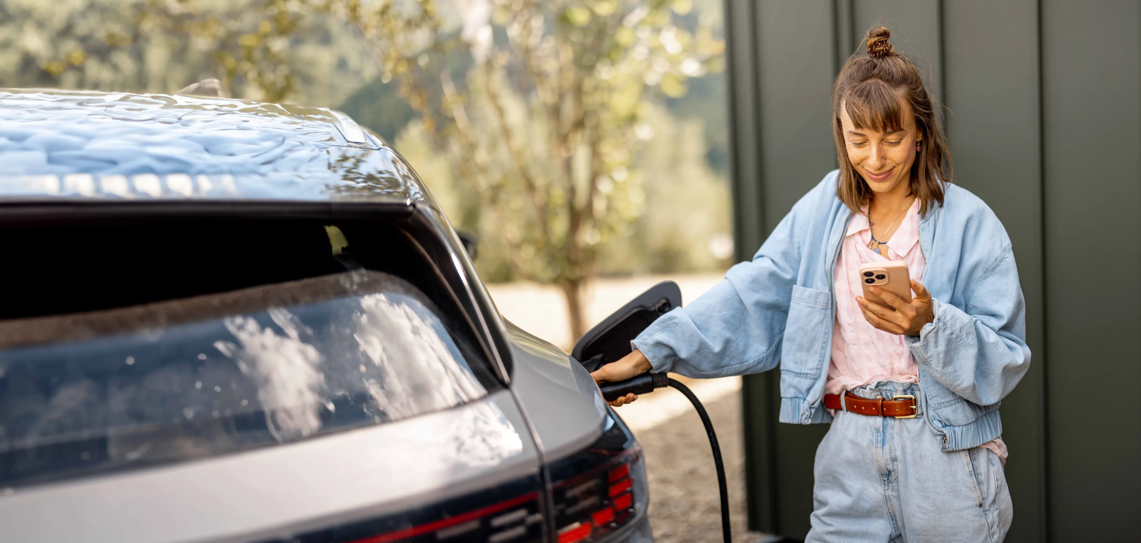 Eine Frau in Jeansjacke lädt ein Elektroauto auf, während sie an einem sonnigen Tag im Freien auf ihr Handy schaut.