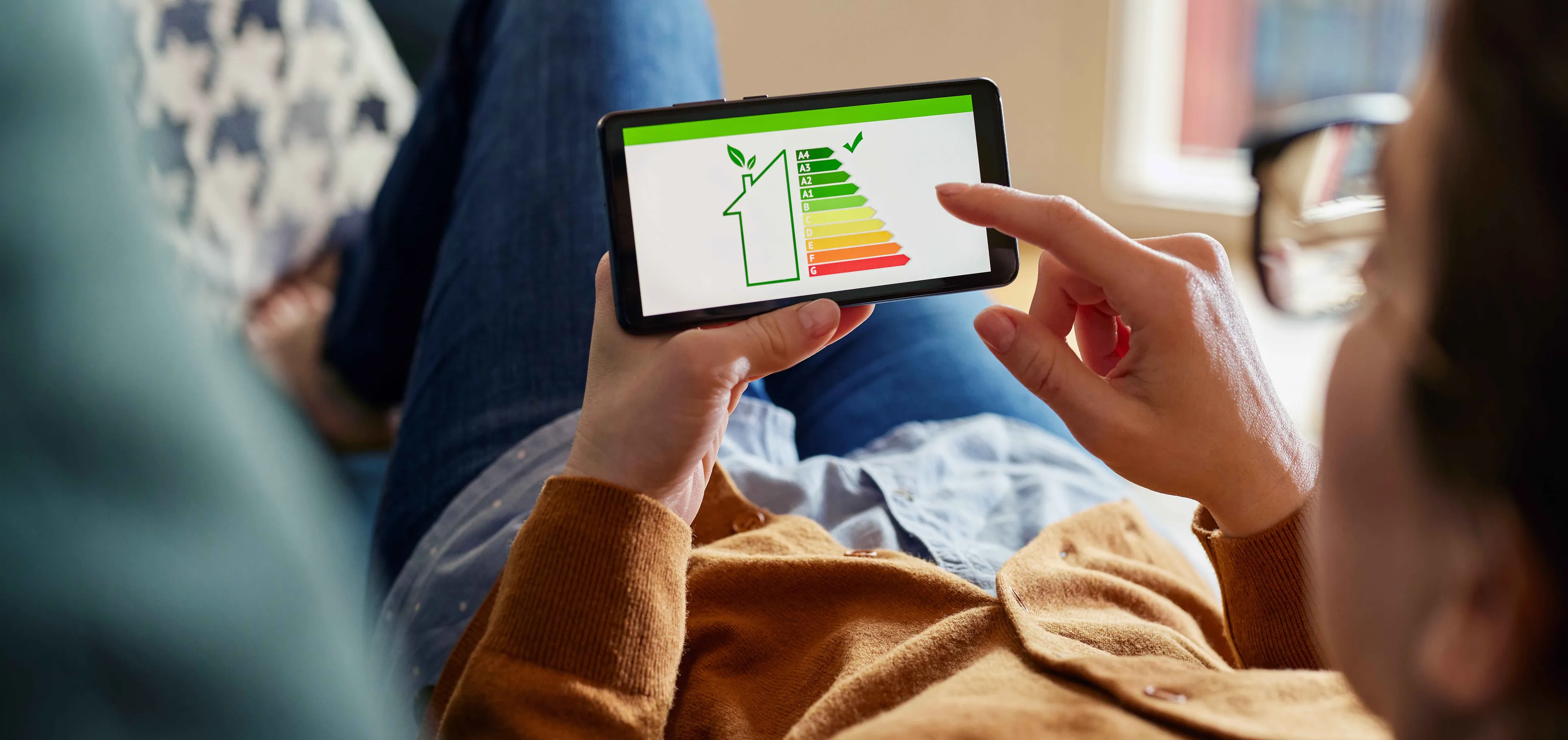 Eine Person hält ein Smartphone in der Hand, auf dem ein Diagramm zur Energieeffizienz mit farbigen Balken und einem Haussymbol angezeigt wird, während sie sich auf einer Couch entspannt.