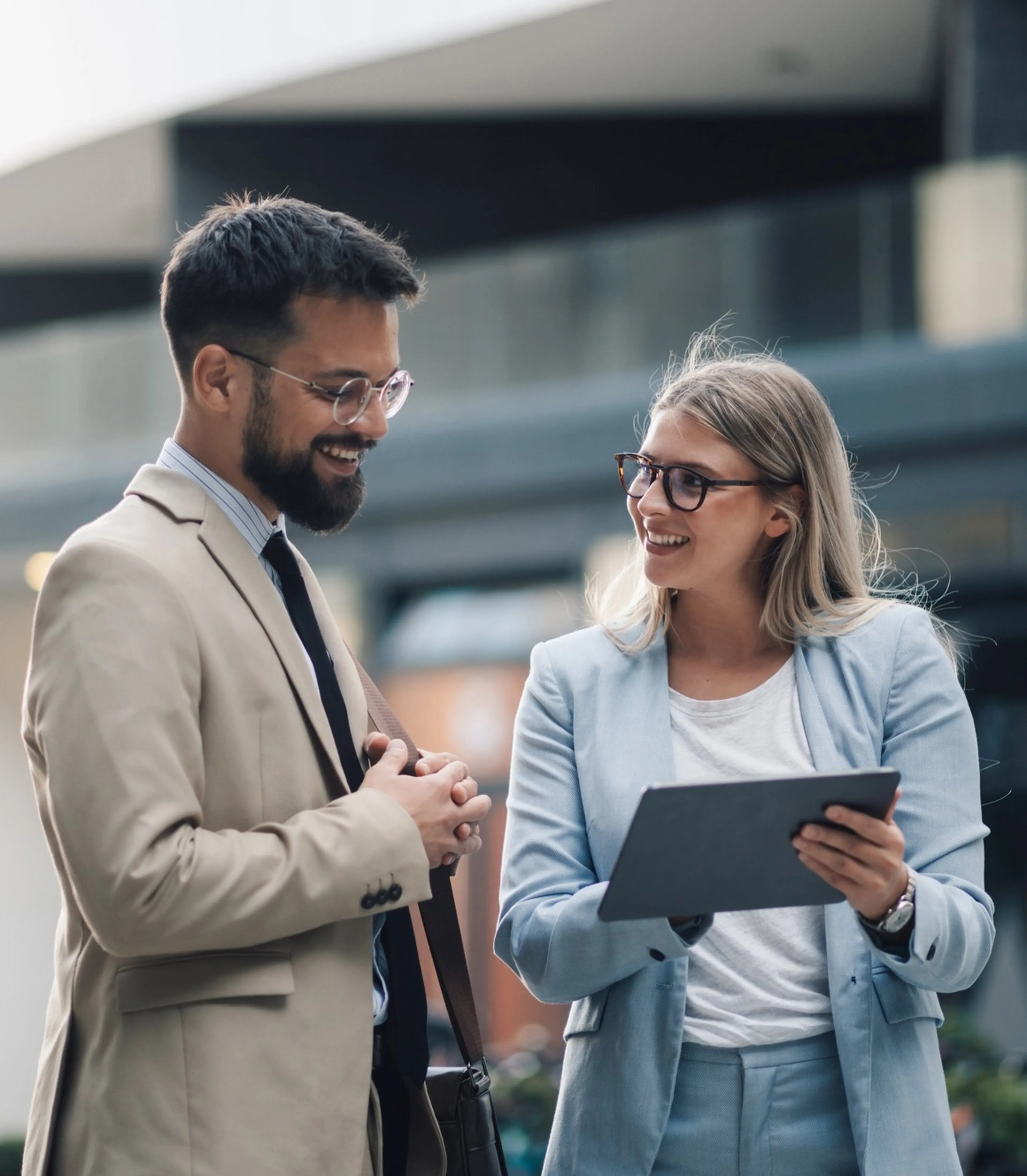 Zwei Berufstätige in Business-Kleidung unterhalten sich im Freien. Einer hält ein Tablet, und beide lächeln. Modernes Gebäude im Hintergrund.