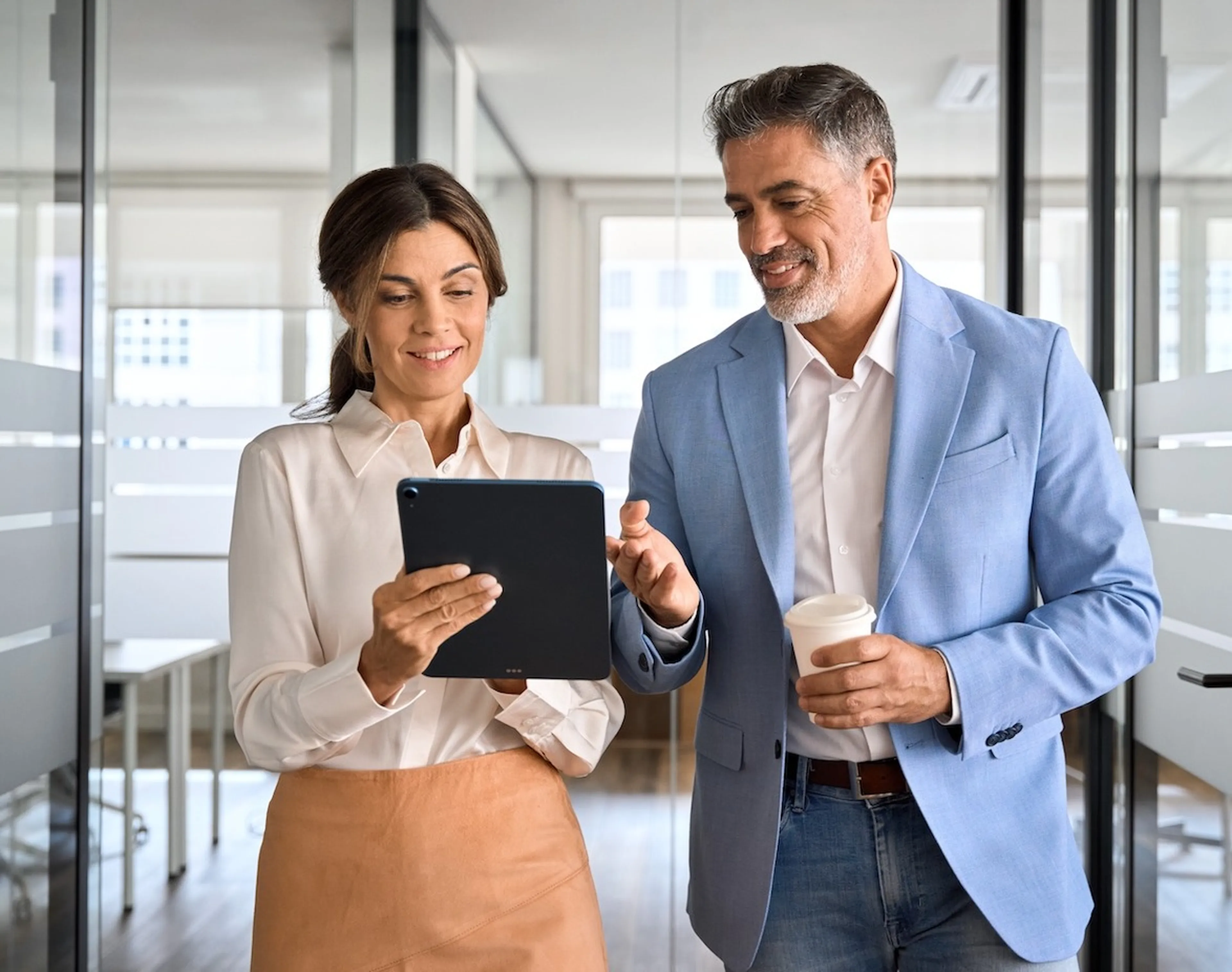 Eine Geschäftsfrau und ein Geschäftsmann laufen durch ein Büro, unterhalten sich und schauen sich etwas am tablet an.