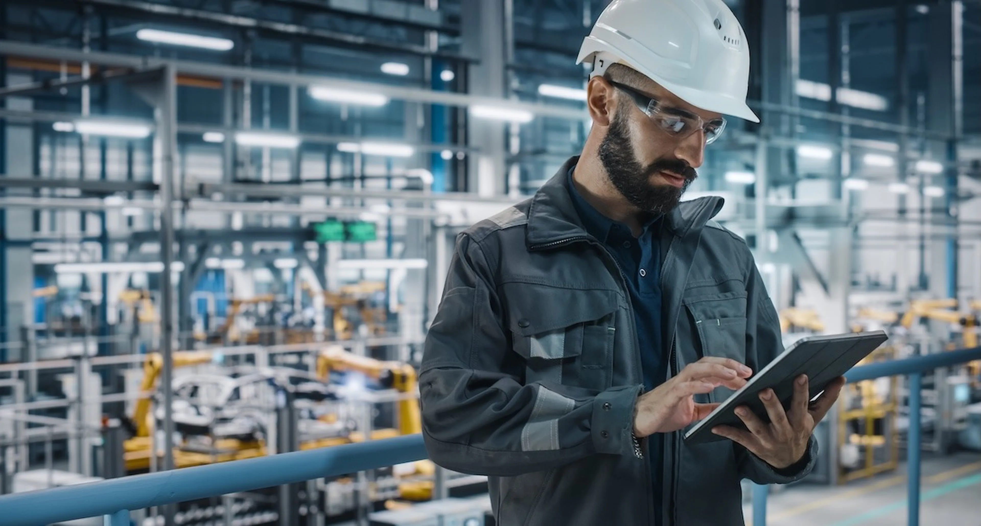 Mann mit Helm und Tablet steht in einer Industriehalle und tippt etwas.