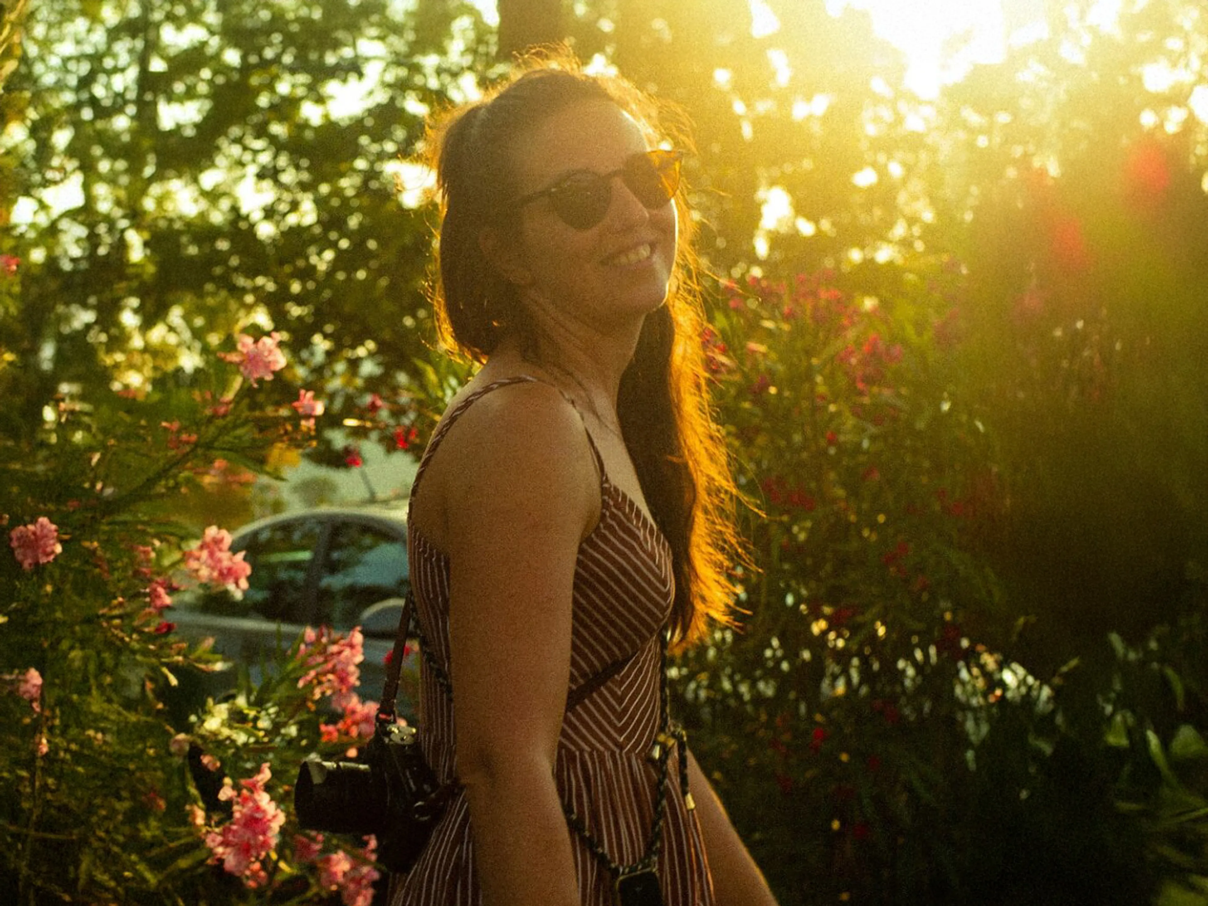 Frau mit Sonnenbrille und Kamera im Sommergarten, umgeben von Blüten, im warmen Sonnenlicht