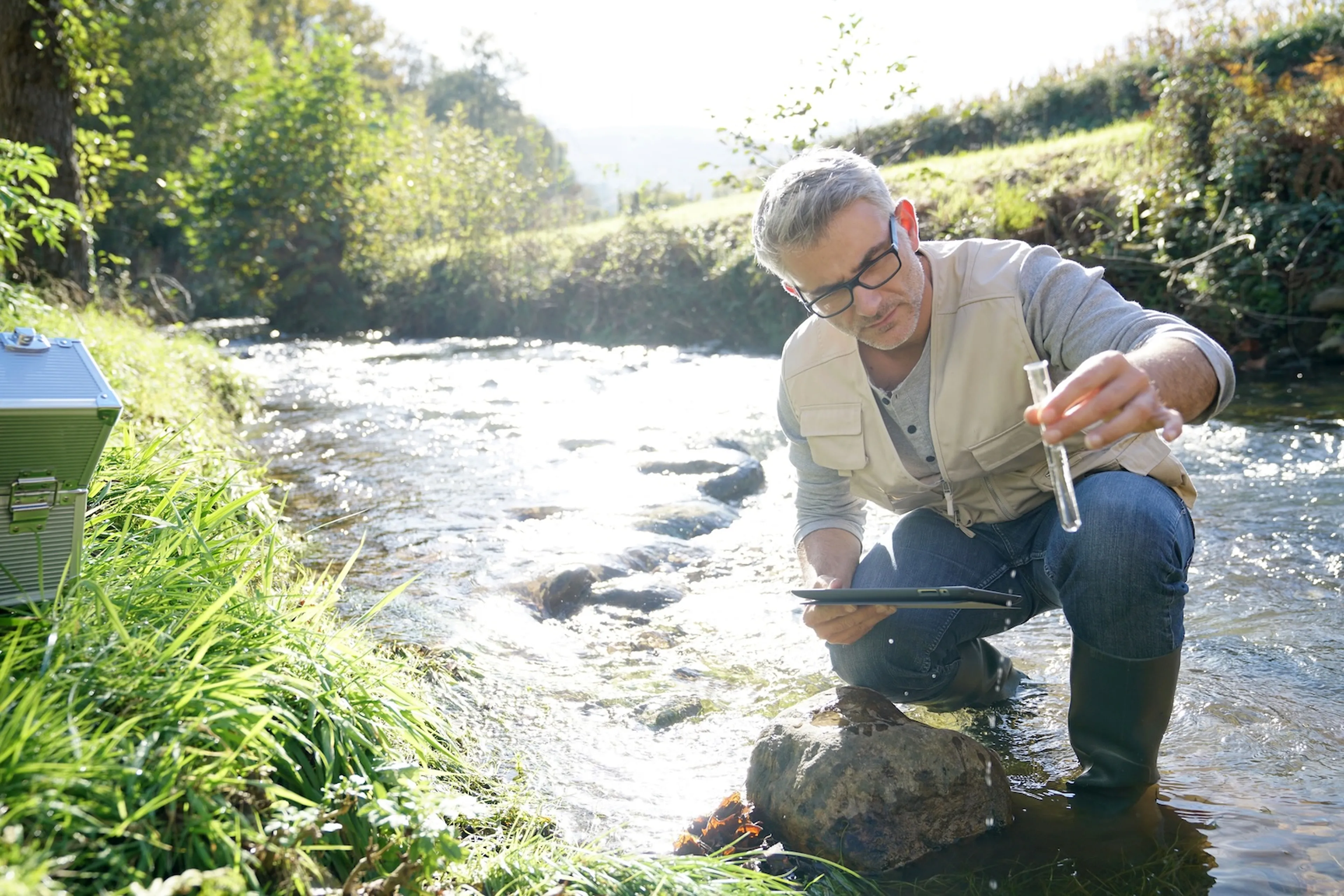 Mann kniet im Fluss und prüft die Wasserqualität mit einem Röhrchen.