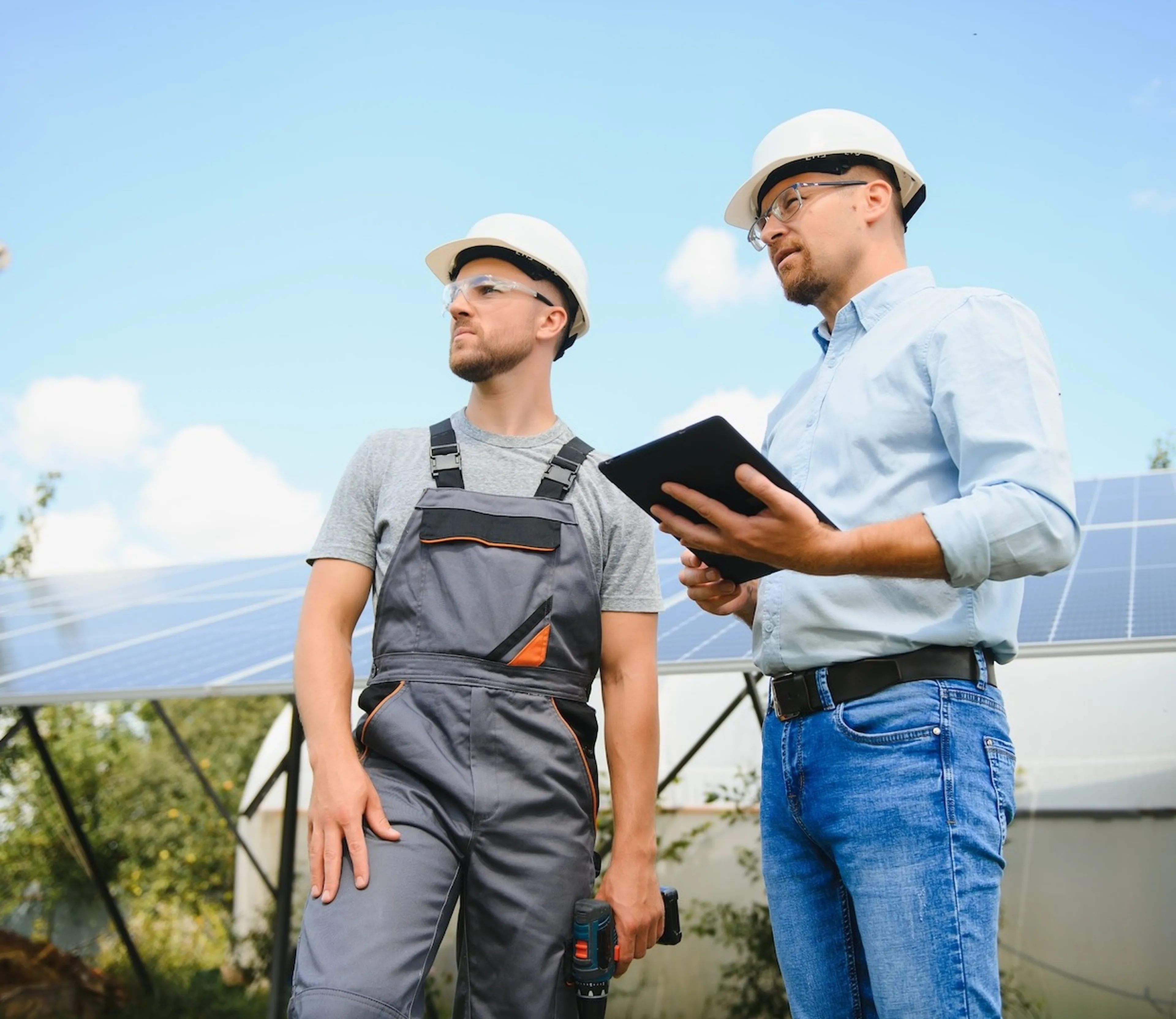 Zwei Männer mit Helmen stehen vor einer Photovoltaikanlage und schauen nach links. Ein Mann hat Bauarbeiterkleidung und eine Bohrmaschine in der Hand, der andere hat ein Tablet in der Hand.