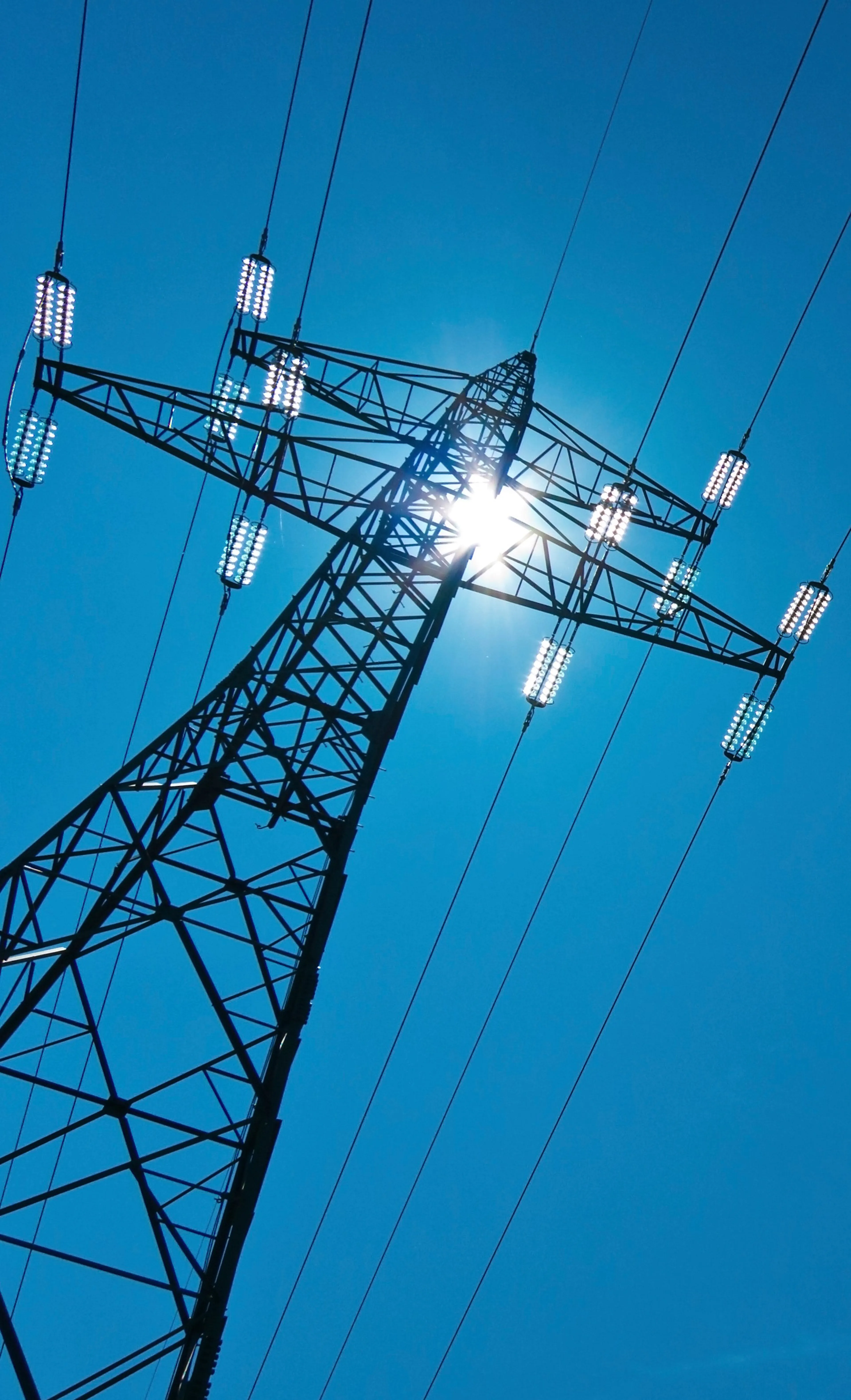 Ein Strommast mit Hochspannungsleitungen ragt vor einem klaren blauen Himmel empor, die Sonne scheint durch das Stahlgerüst.