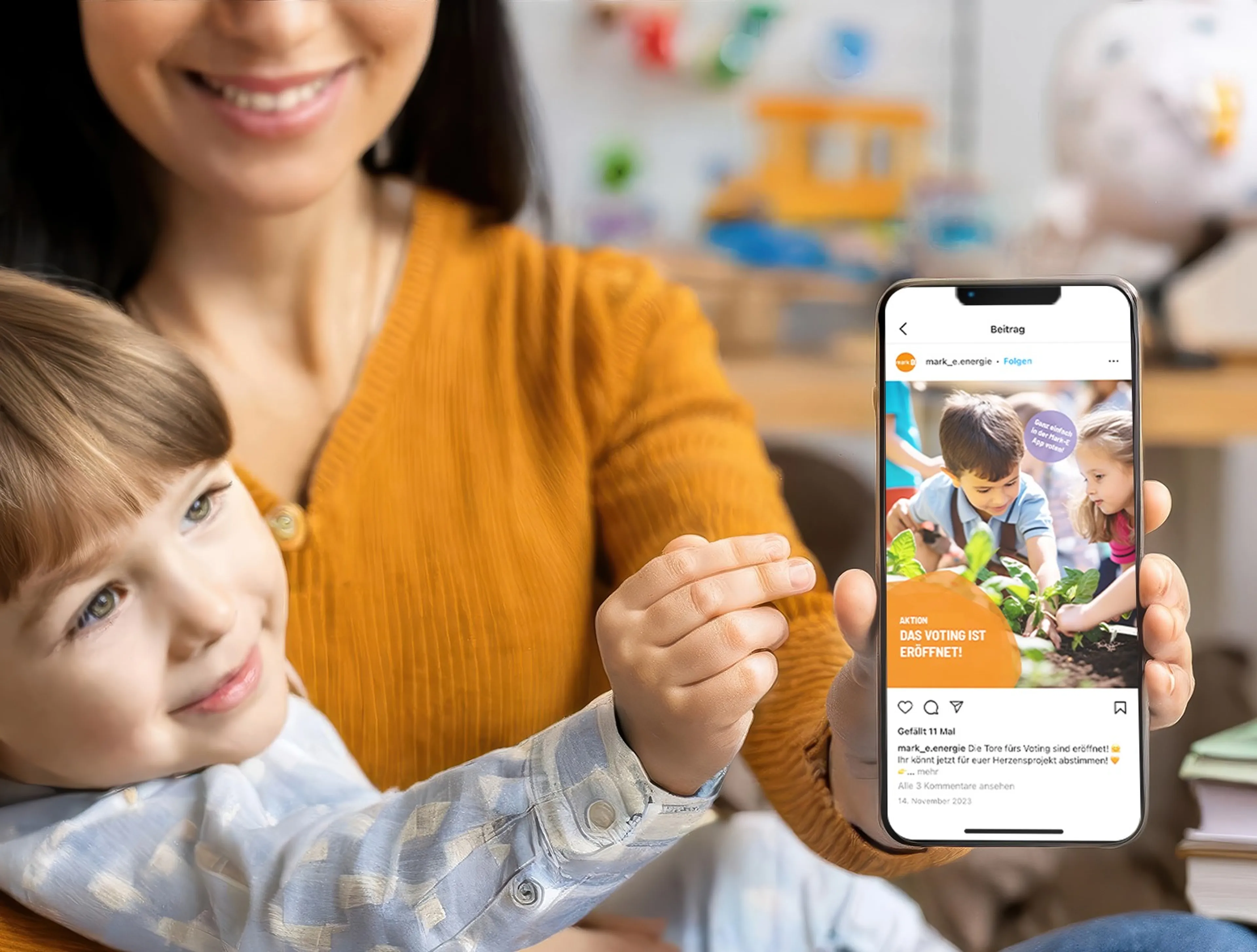 Frau hat auf ihrem Schoß ein Kind und in der einen Hand hält sie ein Smartphone. Die beiden lachen. Auf dem Smartphone zu sehen ist ein Instagram Beitrag, dass das Voting eröffnet ist.