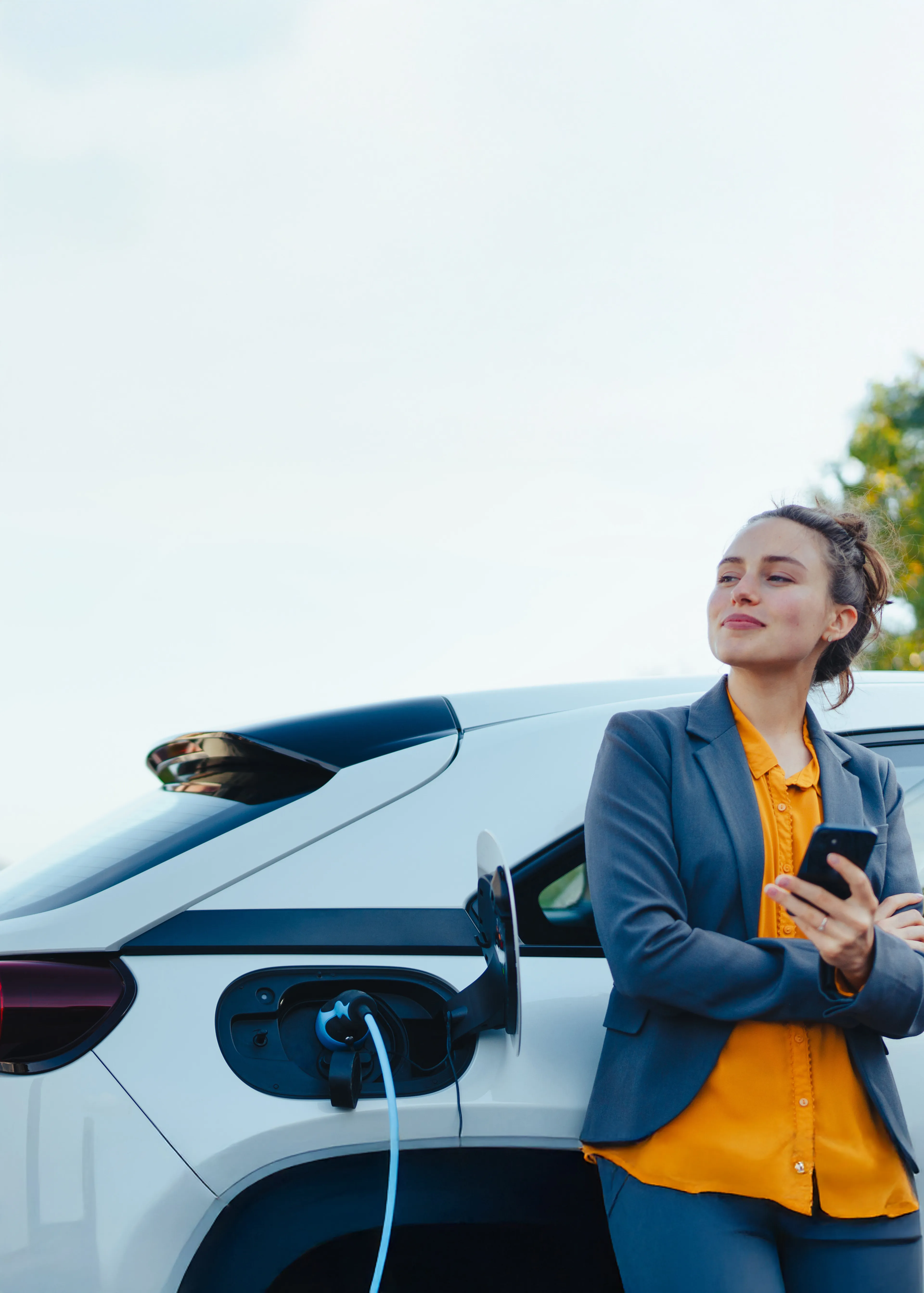 Frau blickt in die Ferne, während sie mit einem Smartphone am ladenden Elektroauto steht