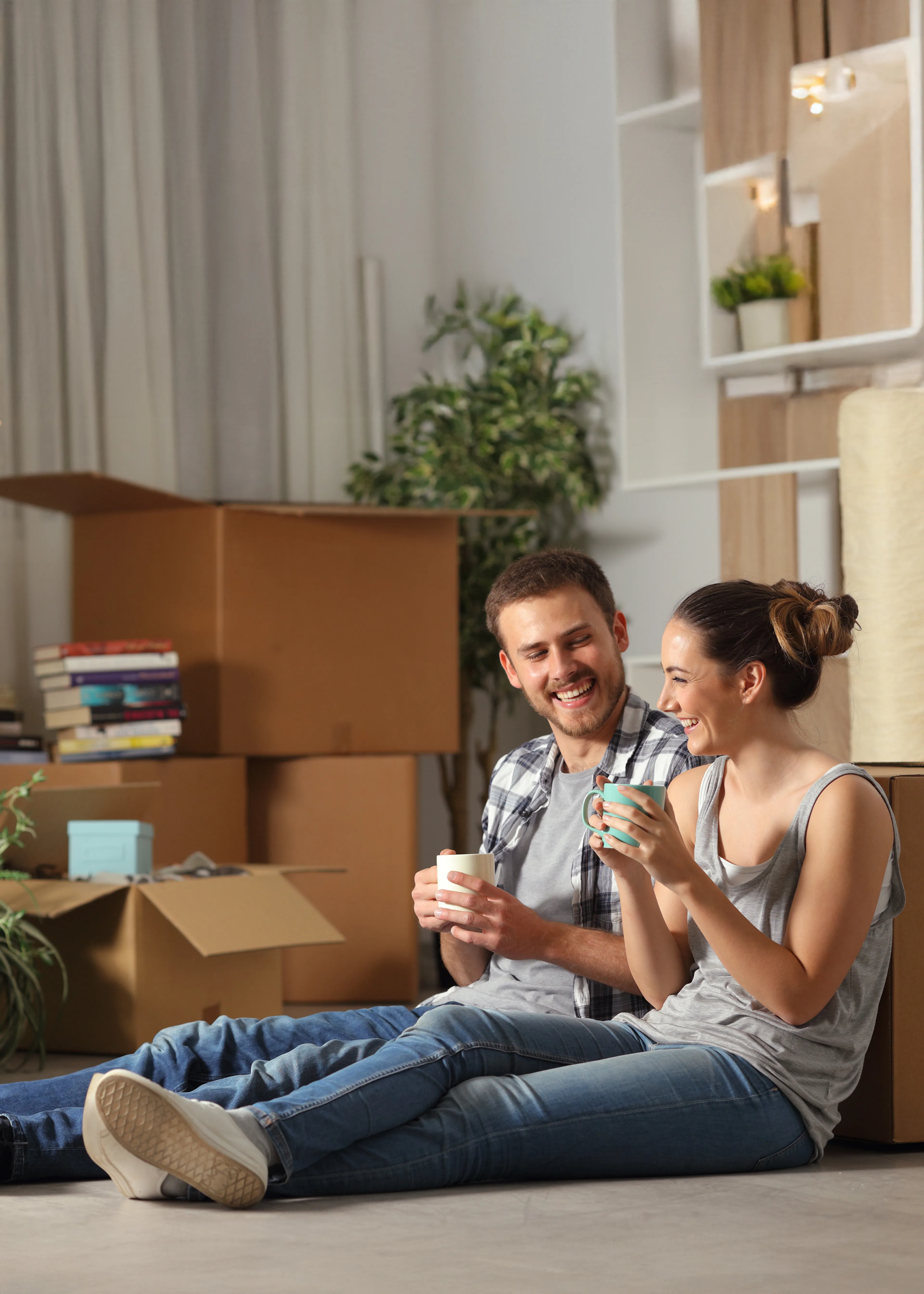 Junges Paar sitzt lachend mit Tassen zwischen Umzugskartons in der neuen Wohnung