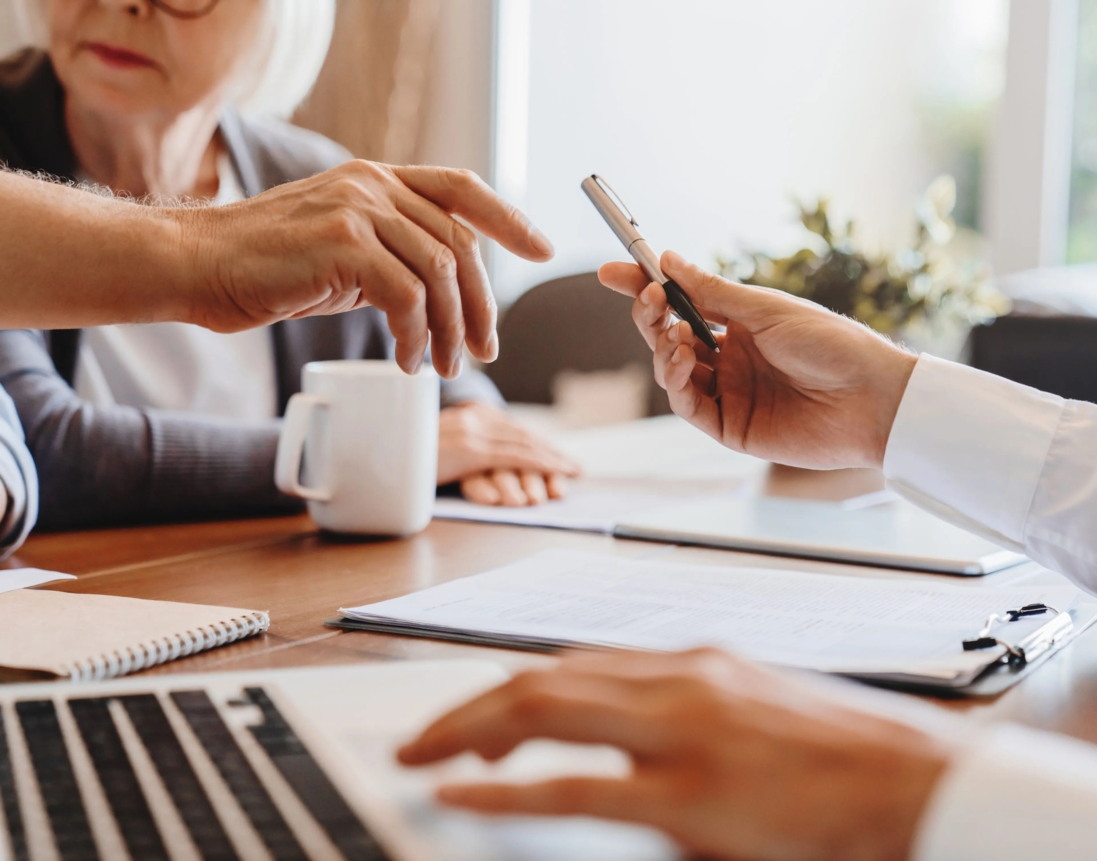 Auf einem Tisch liegen viele Dokumente. Drei Menschen sitzen am Tisch und die eine Person reicht einer anderen einen Stift.