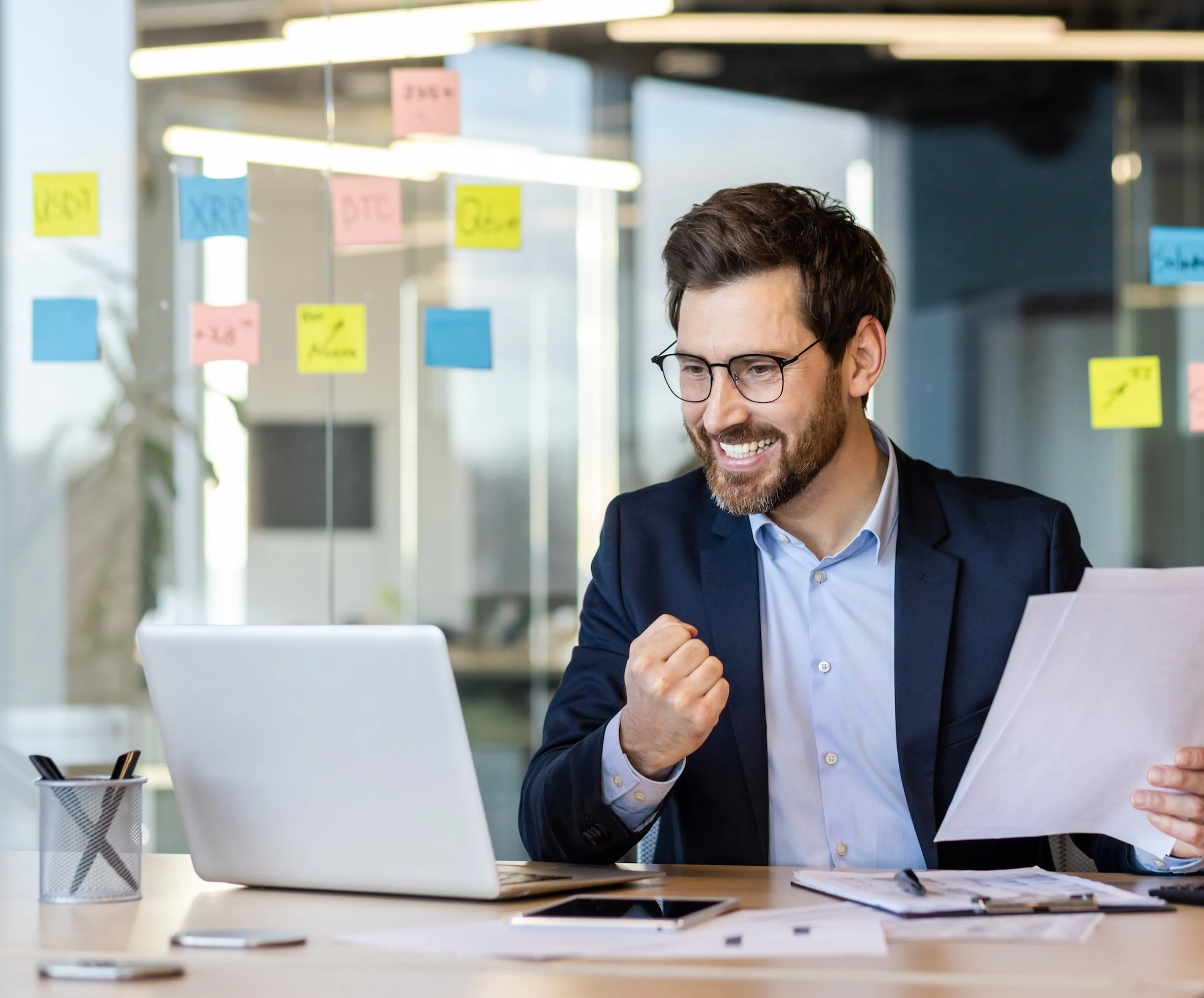 Geschäftsmann freut sich über positive Ergebnisse bei der Arbeit am Laptop, bunte Post-its im Hintergrund.