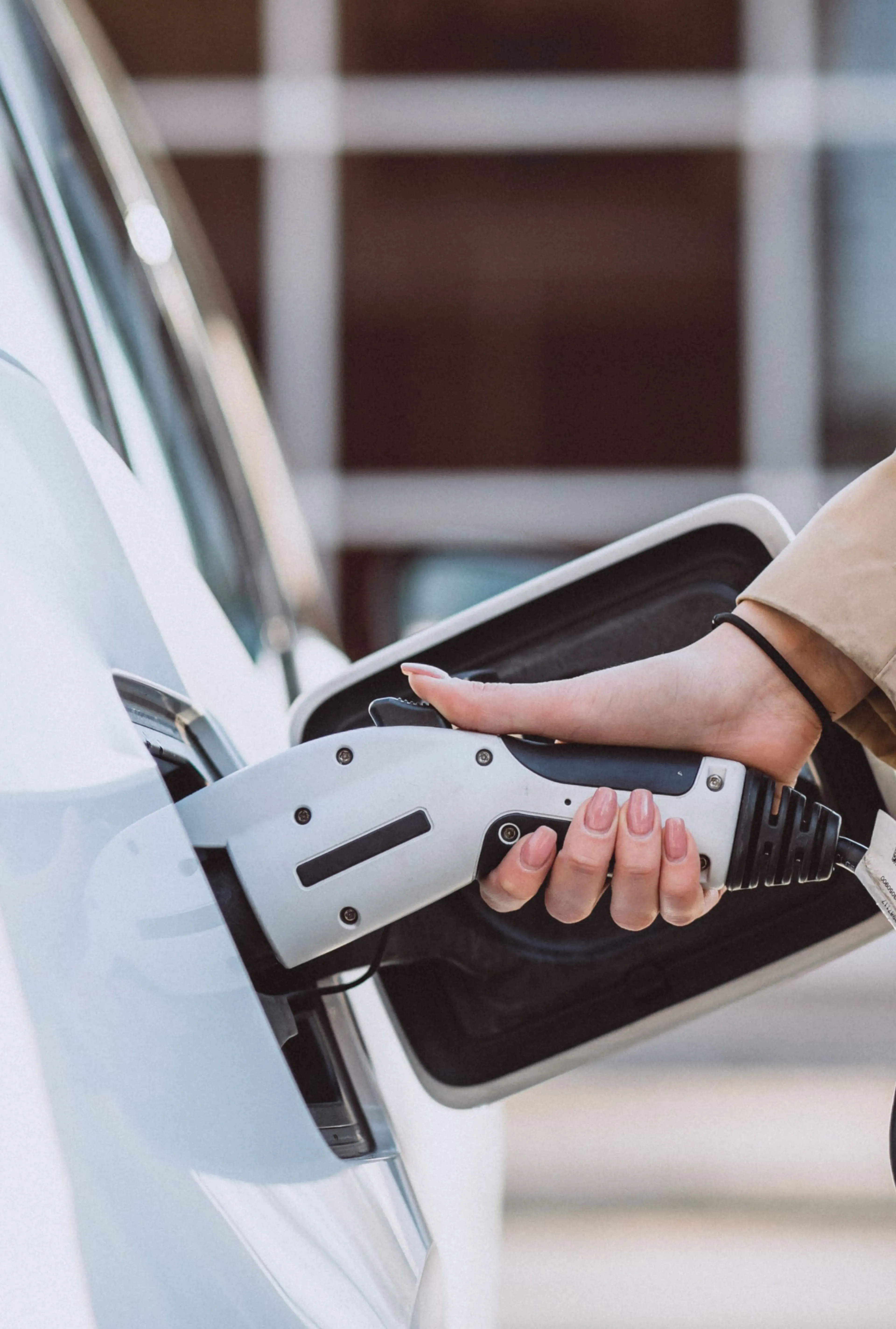Person, die ein Ladegerät an ein Elektroauto anschließt, Nahaufnahme von Hand und Ladeanschluss, mit einem unscharfen Gebäude im Hintergrund.