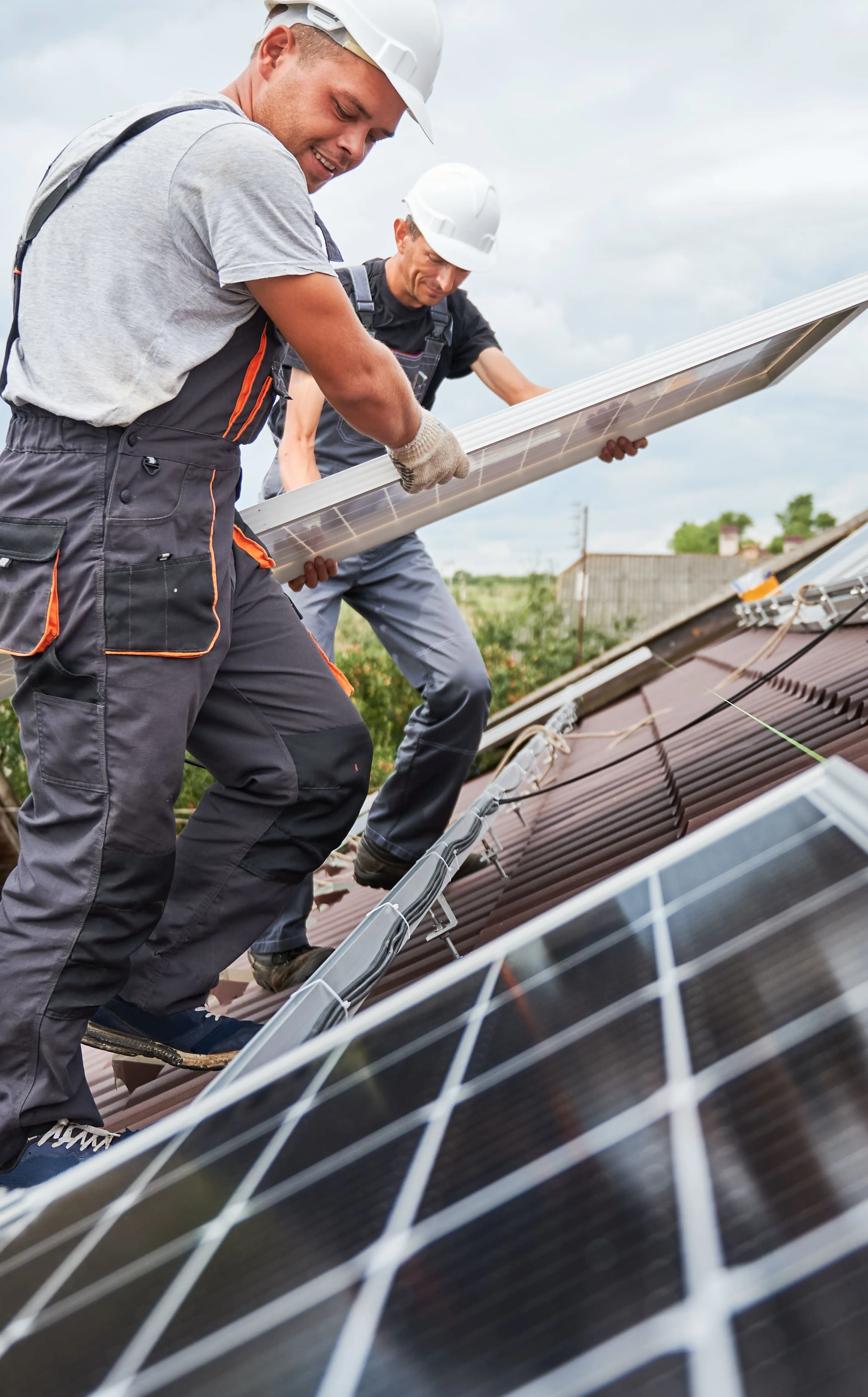 Zwei Arbeiter mit Schutzhelmen bei der Installation von Solarmodulen auf einem Schrägdach, mit einem bewölkten Himmel im Hintergrund.