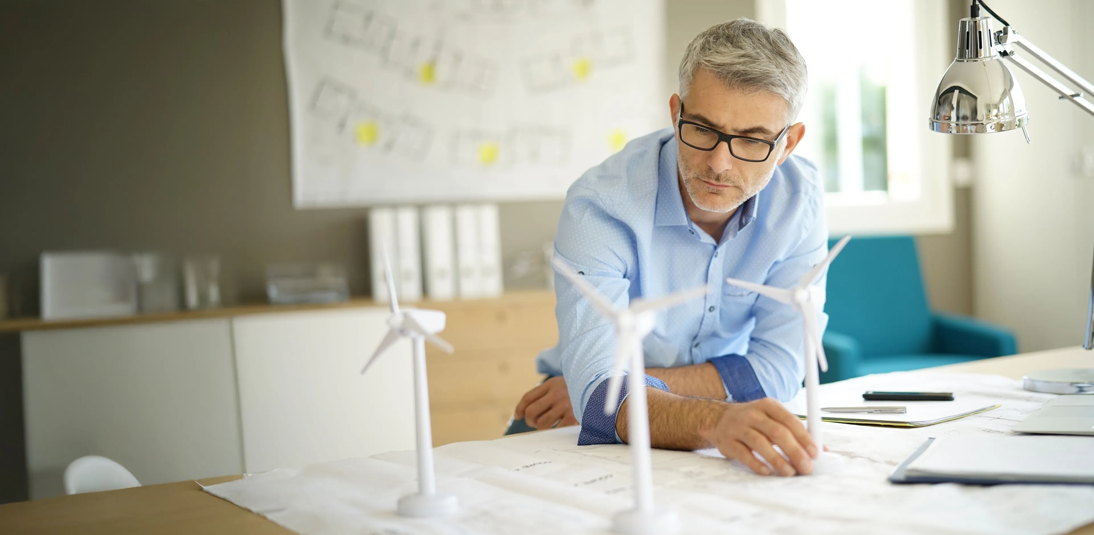 Mann sitzt am Schreibtisch und schaut sich Dokumente und Modelle von Windrädern an.