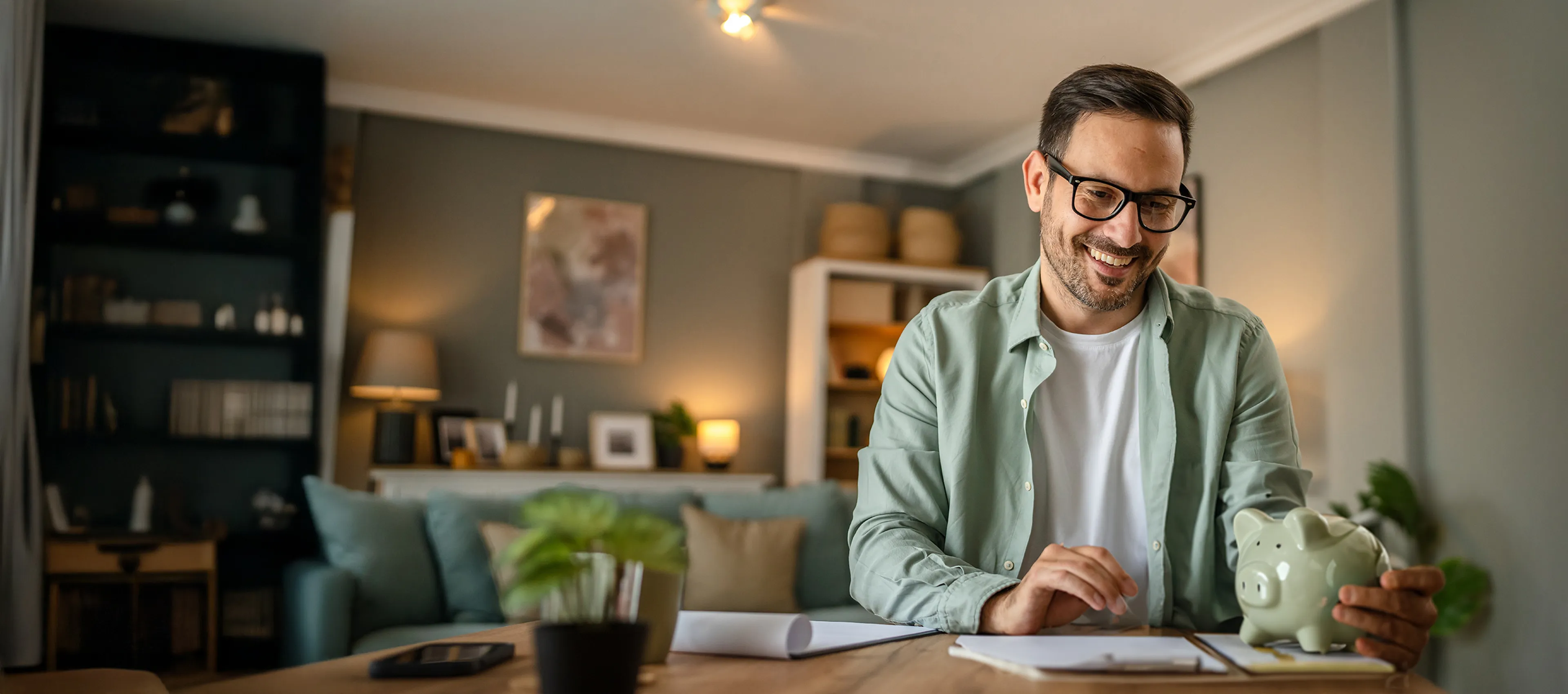 Mann sitzt an einem Tisch im Wohnzimmer, auf dem Dokumente liegen. Er hält ein Sparschwein in der Hand.