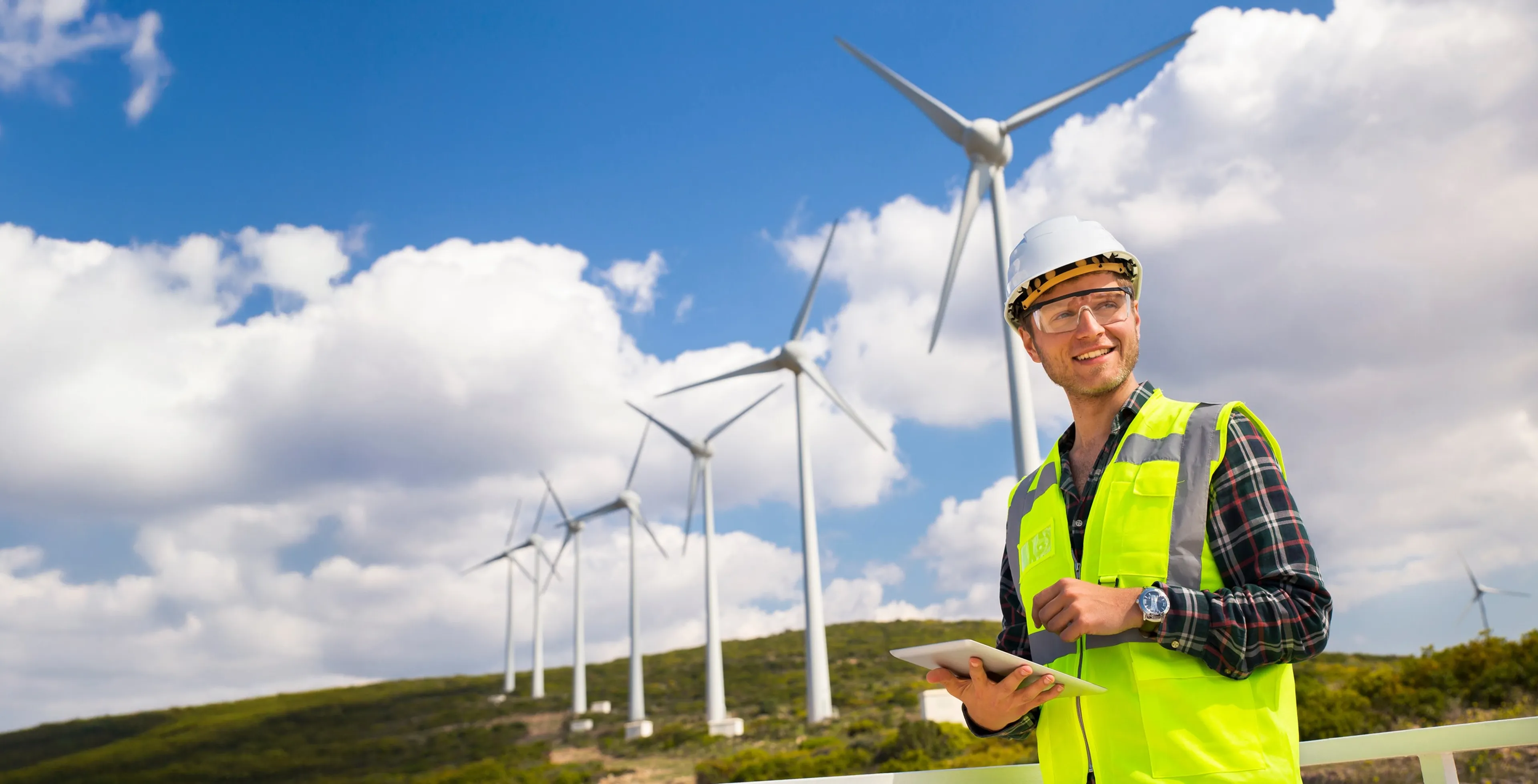 Techniker steht im Vordergrund und lächelt. Im Hintergrund ist ein Hügel mit mehreren Windrädern.