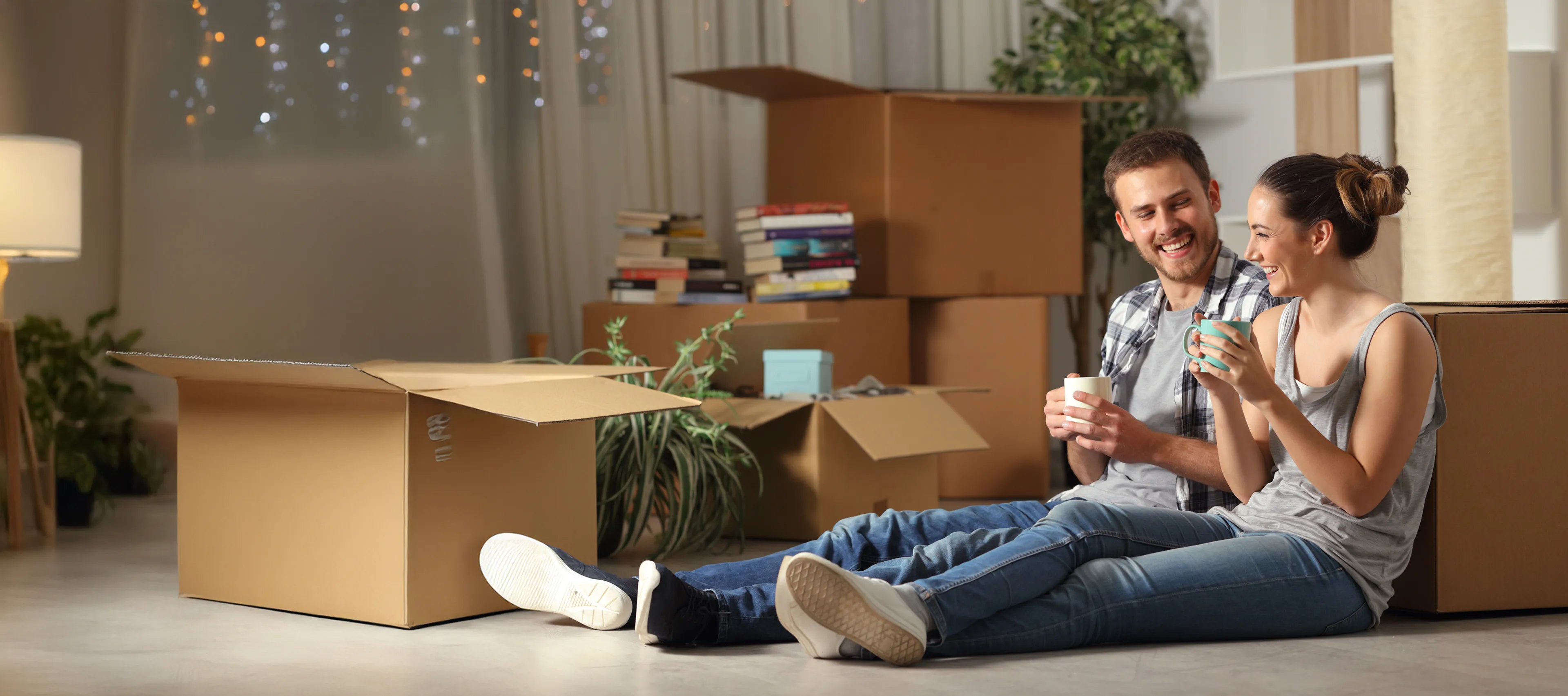 Junges Paar sitzt lachend mit Tassen zwischen Umzugskartons in der neuen Wohnung