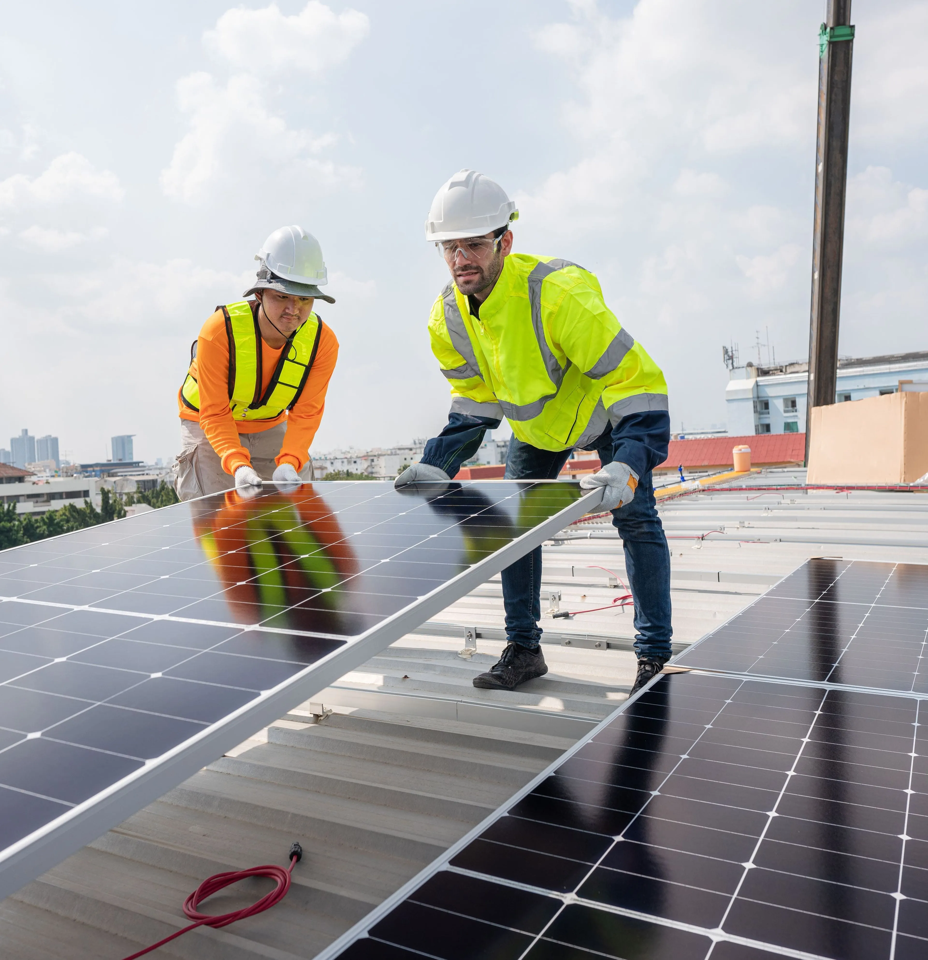 Zwei Arbeiter in Schutzkleidung installieren bei bewölktem Himmel Solarmodule auf einem Dach, mit städtischen Gebäuden im Hintergrund.
