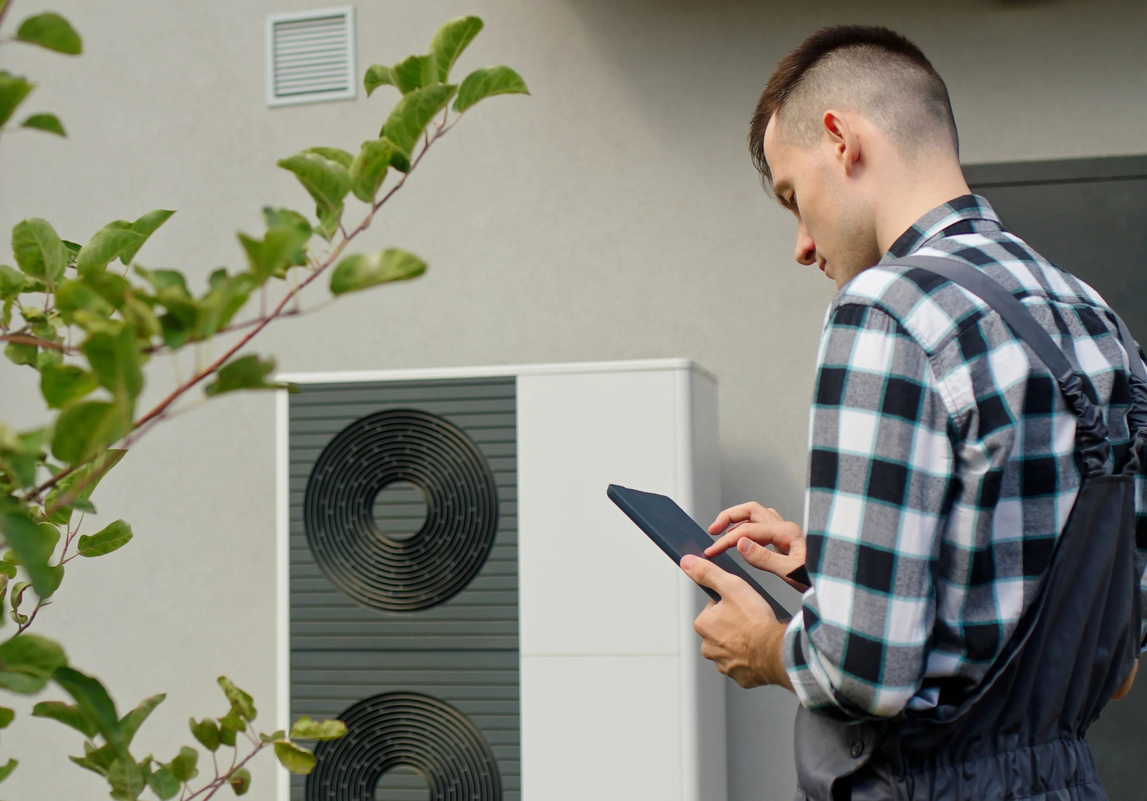 Techniker prüft Wärmepumpe an der Außenwand eines Hauses mit Tablet