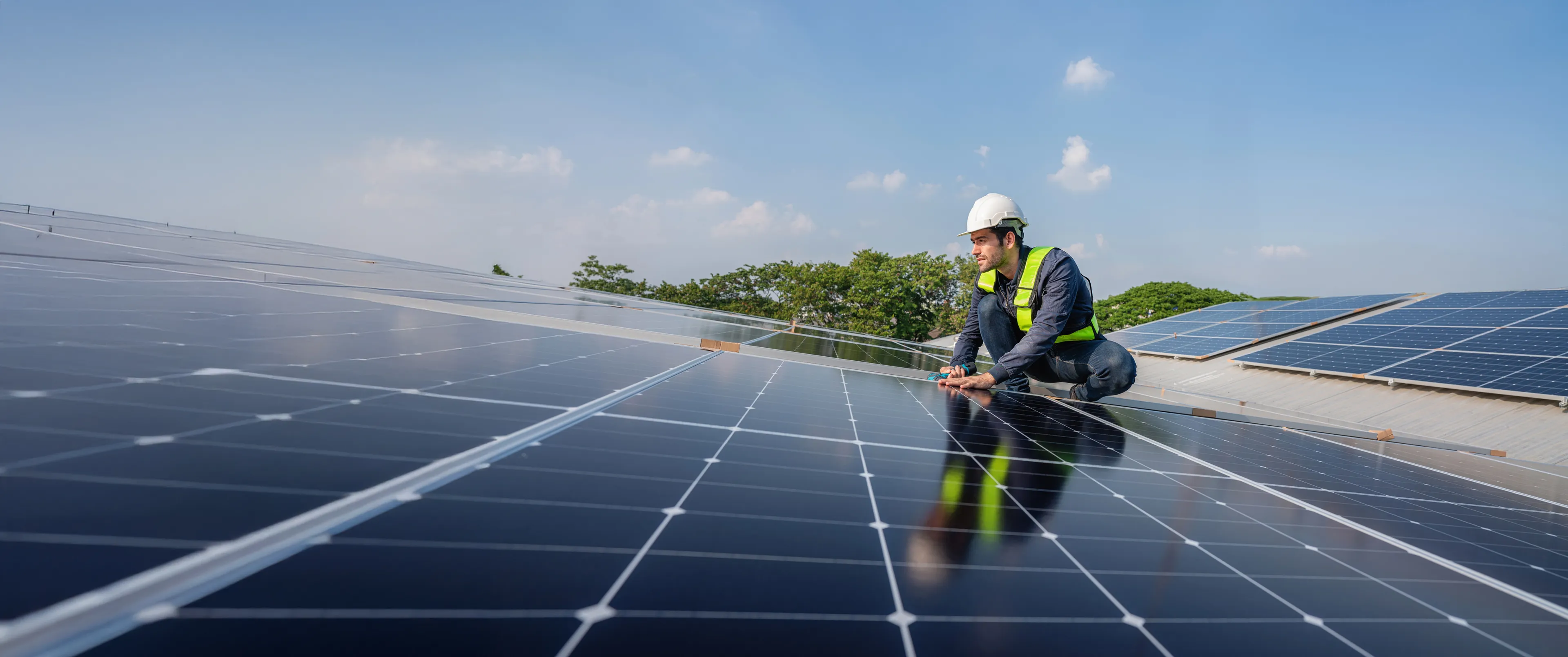 Ein Arbeiter mit Schutzweste und Helm installiert bei strahlend blauem Himmel Solarzellen auf einem Dach.