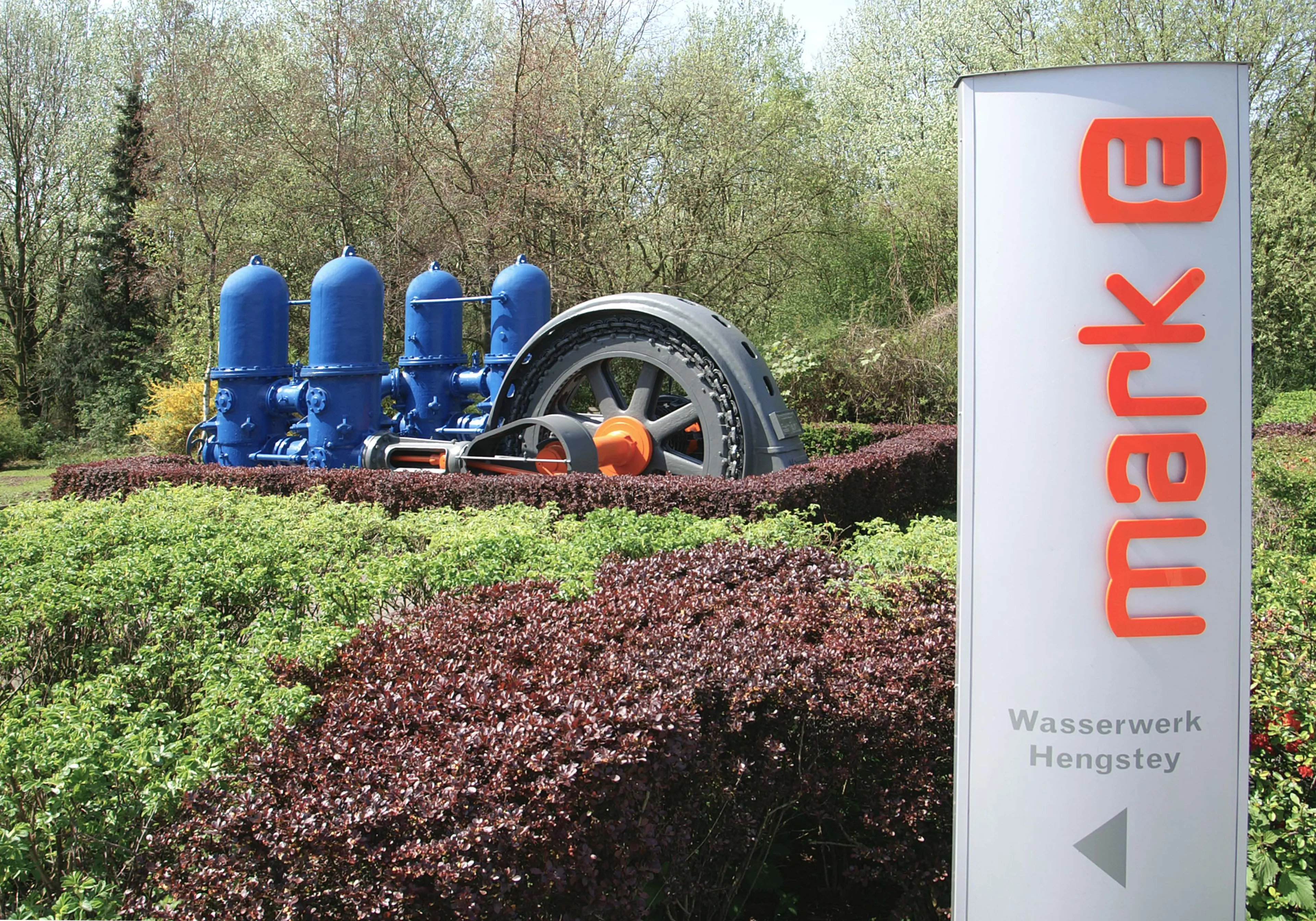 Historische Industrieanlage mit Schwungrad, im Vordergrund ein Schild mit dem Logo „mark E“ und dem Hinweis „Wasserwerk Hengstey“.