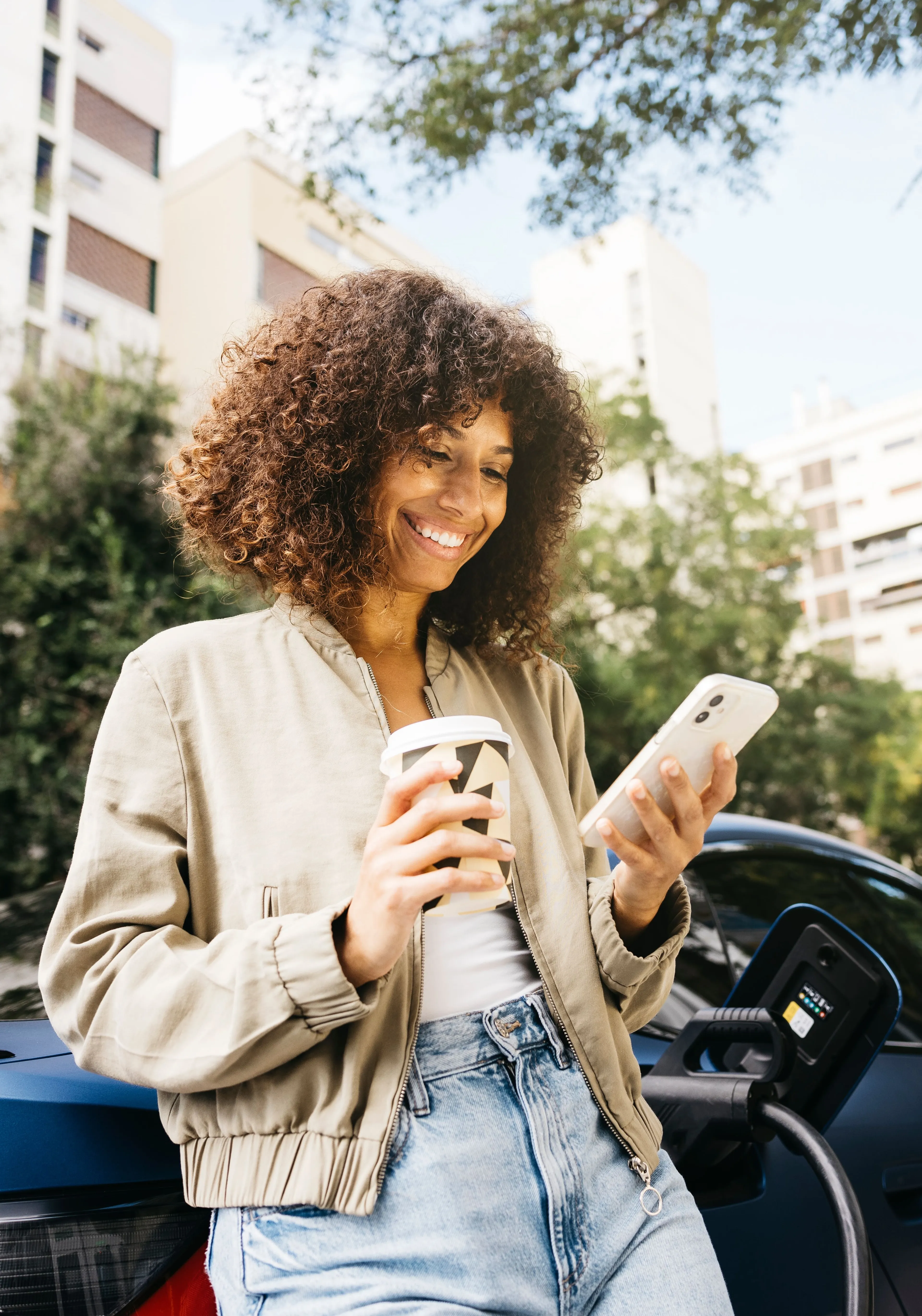 Eine Frau mit lockigem Haar lehnt an einem Elektroauto, hält eine Kaffeetasse in der Hand und schaut auf ihr Telefon, in einer von Bäumen gesäumten Straße.