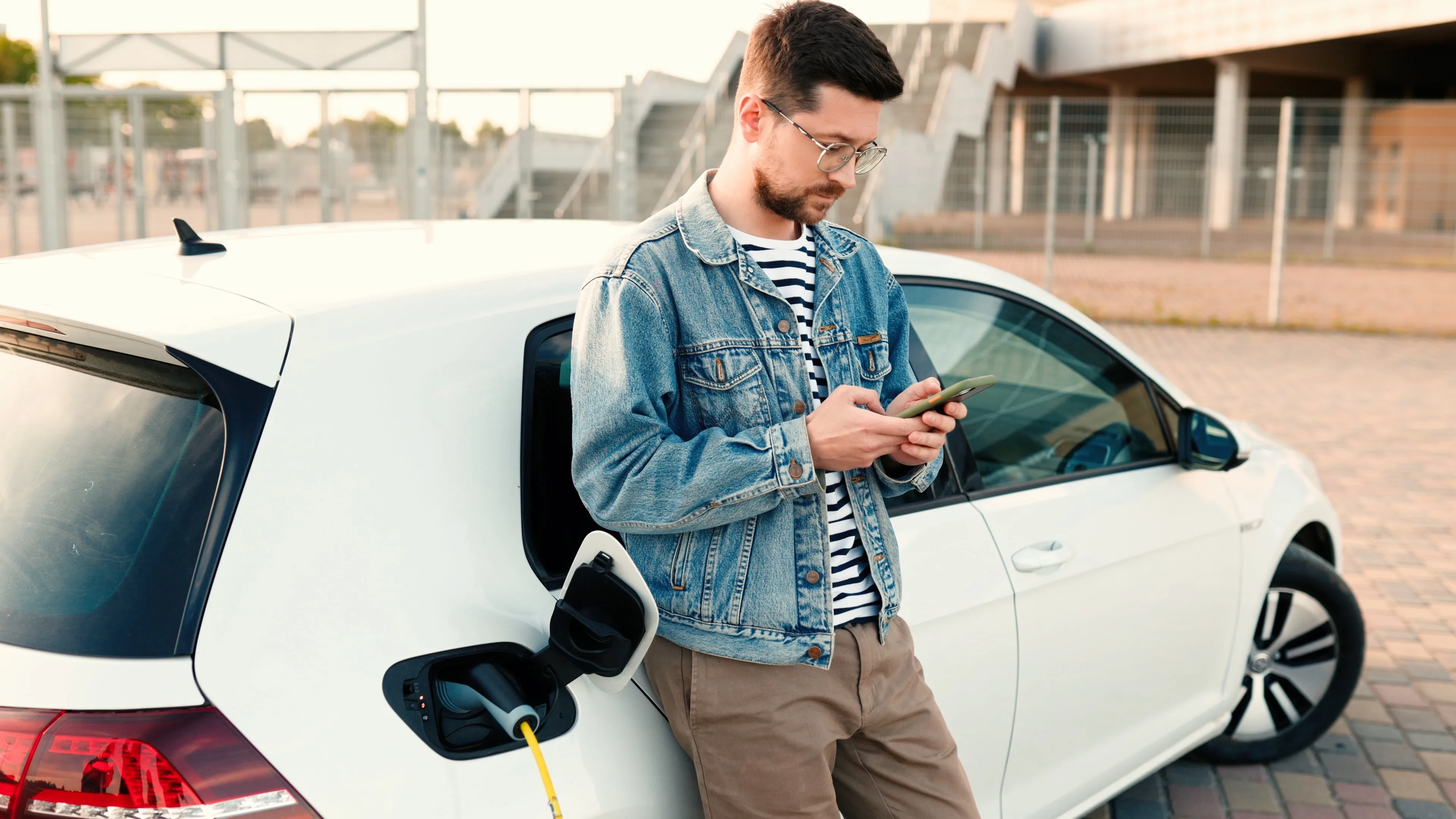 Mann in Jeansjacke benutzt sein Smartphone, während er sich an ein weißes Elektroauto lehnt, das gerade aufgeladen wird.