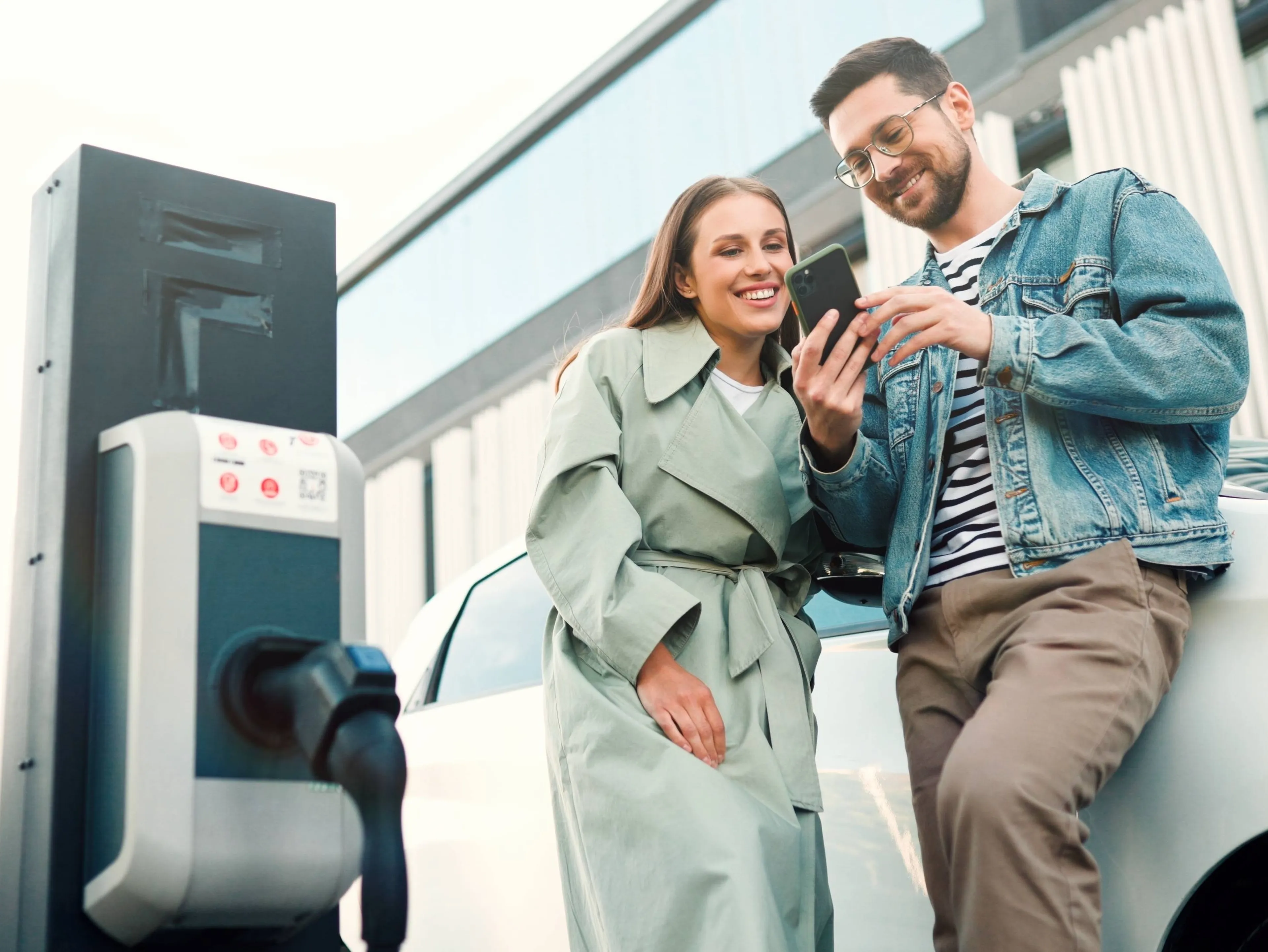 Ein junges Paar steht an einer E-Ladestation, lächelt und betrachtet gemeinsam ein Smartphone. Im Hintergrund ist ihr Elektroauto zu sehen.