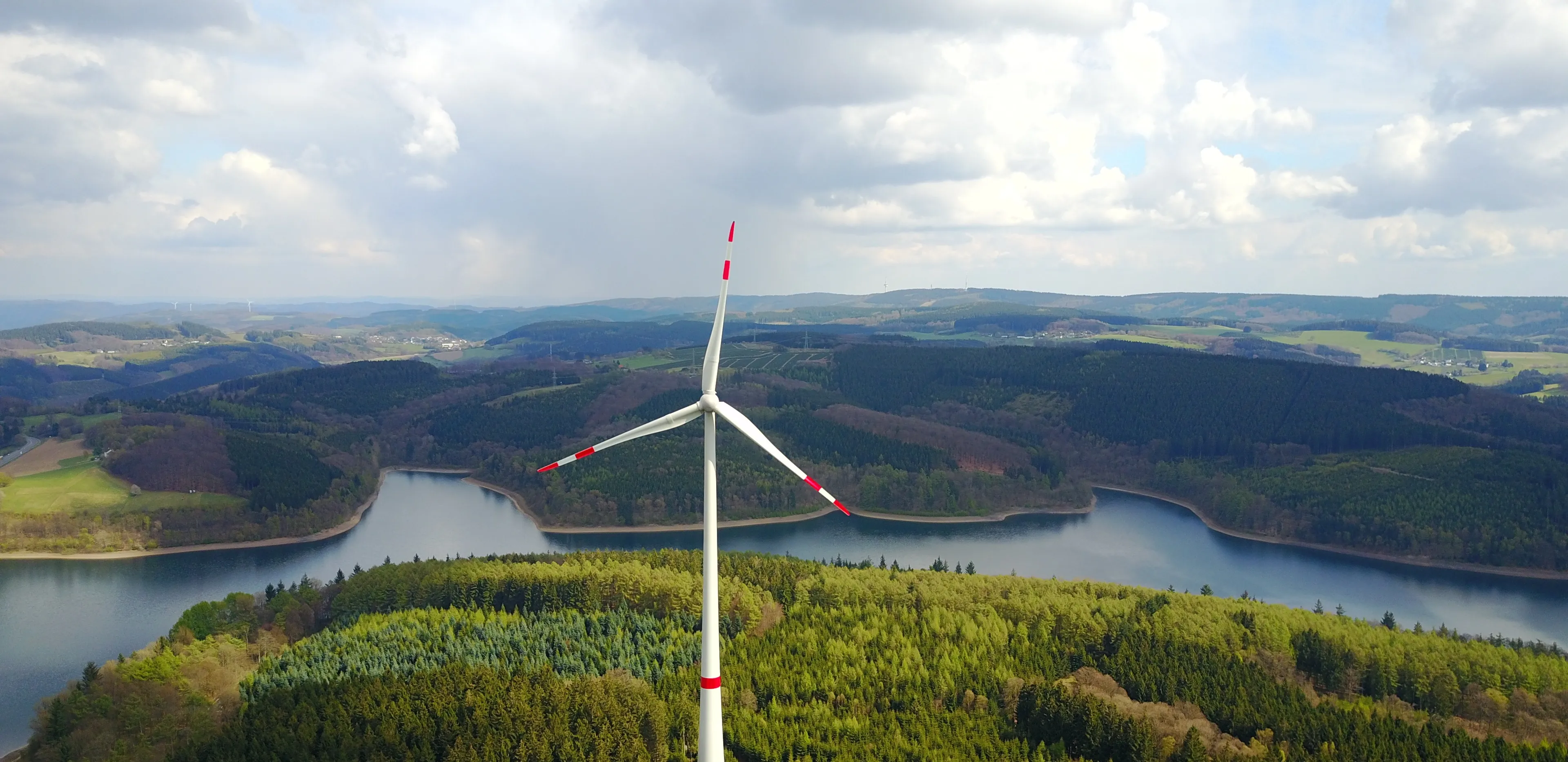 Windkraftwerk im Wald vor einem Fluss mit Bergen im Hintergrund