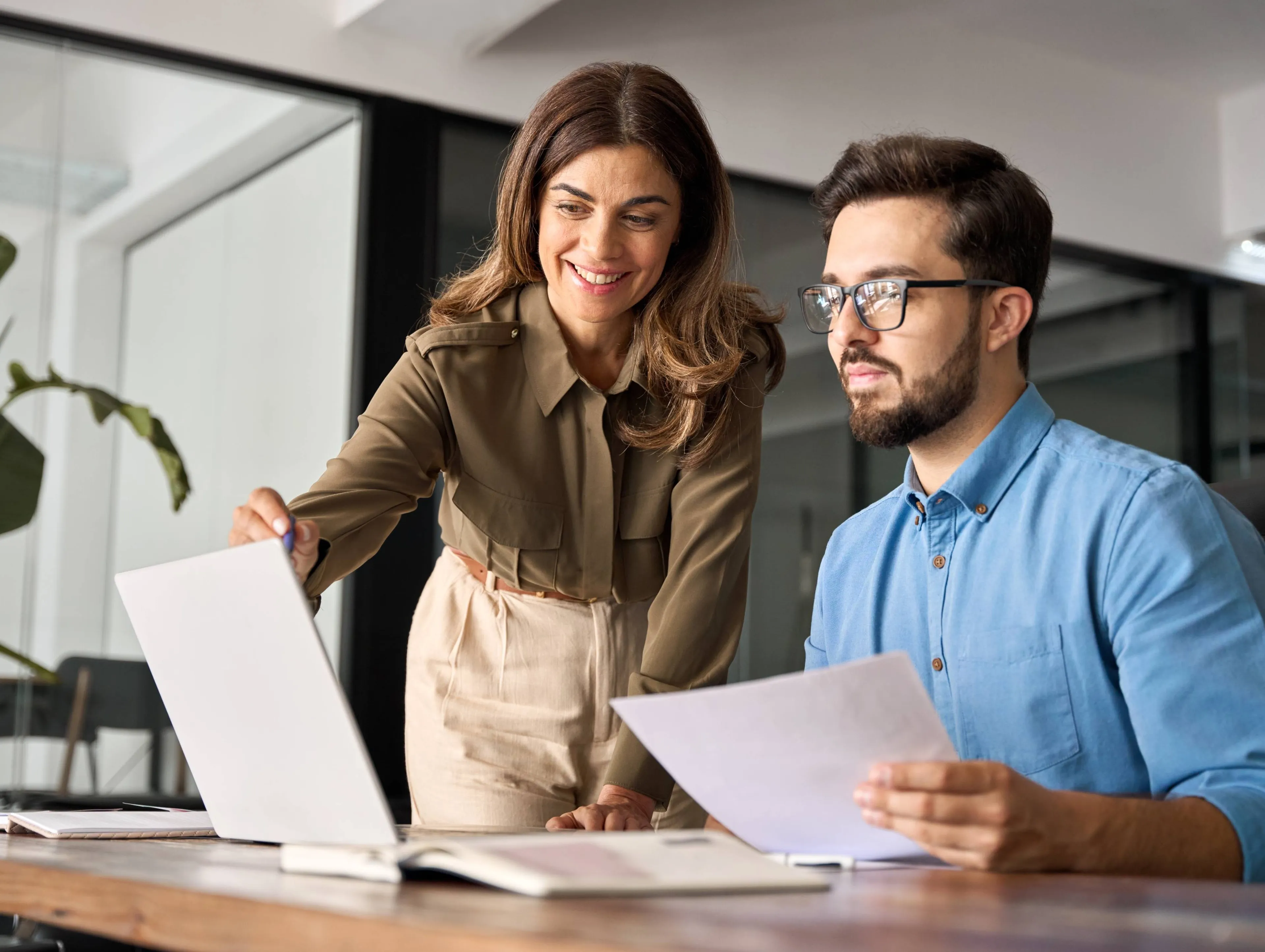Zwei Personen in einem modernen Büro besprechen gemeinsam Informationen am Laptop und mit ausgedruckten Unterlagen.