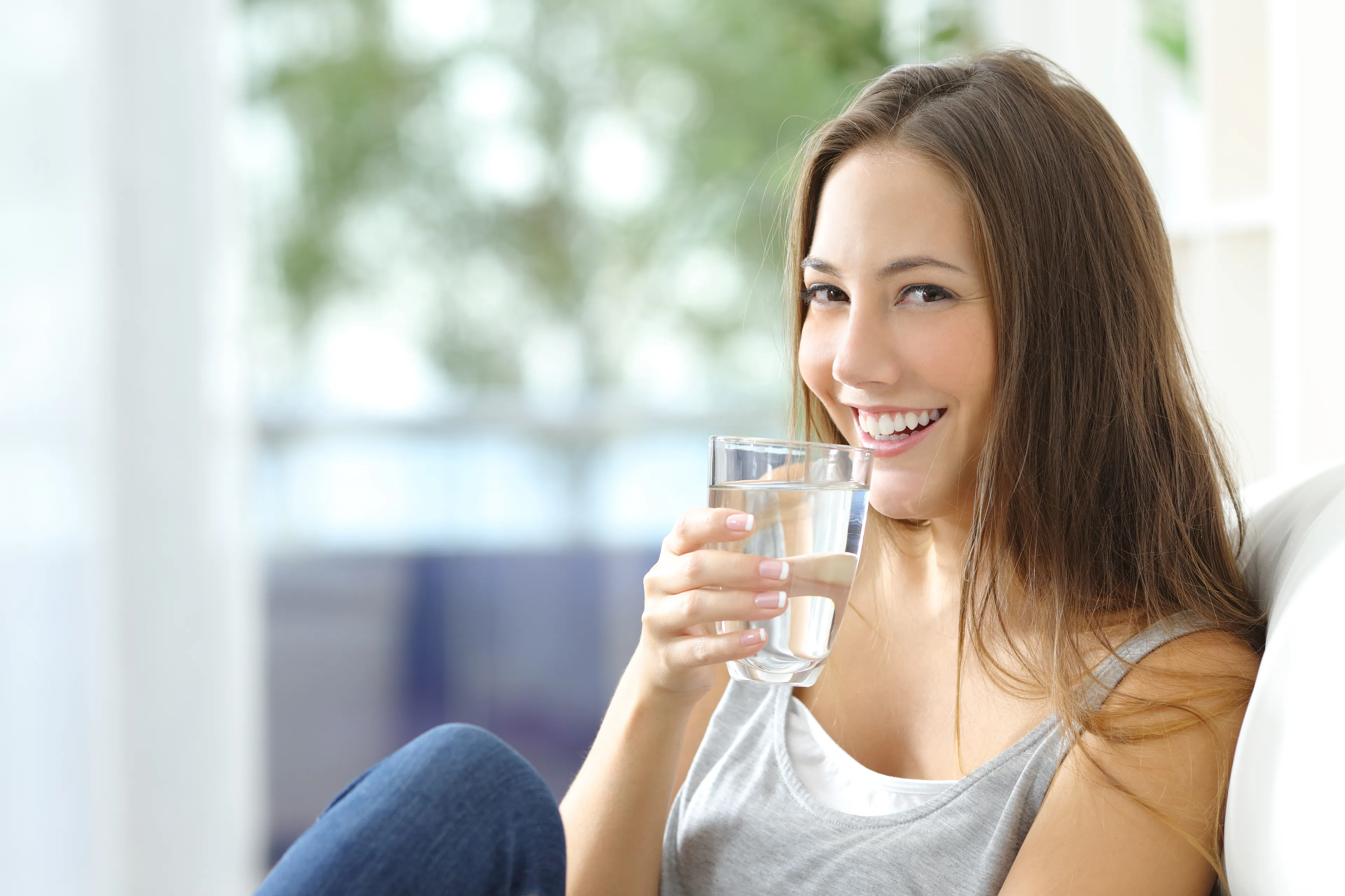 Junge Frau sitzt auf dem Sofa und trinkt ein Glas frisches Trinkwasser