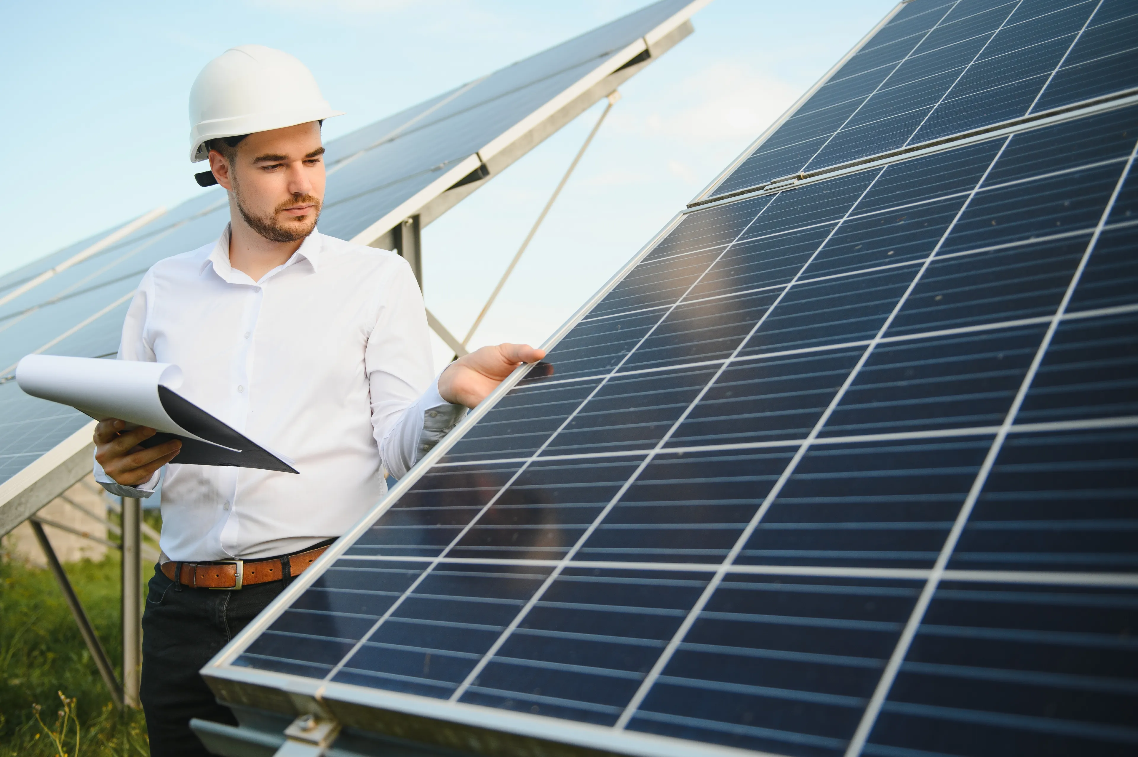 Mann mit weißem Schutzhelm bei der Inspektion von Solarmodulen, mit Dokumenten in der Hand, in einem Solarpark.