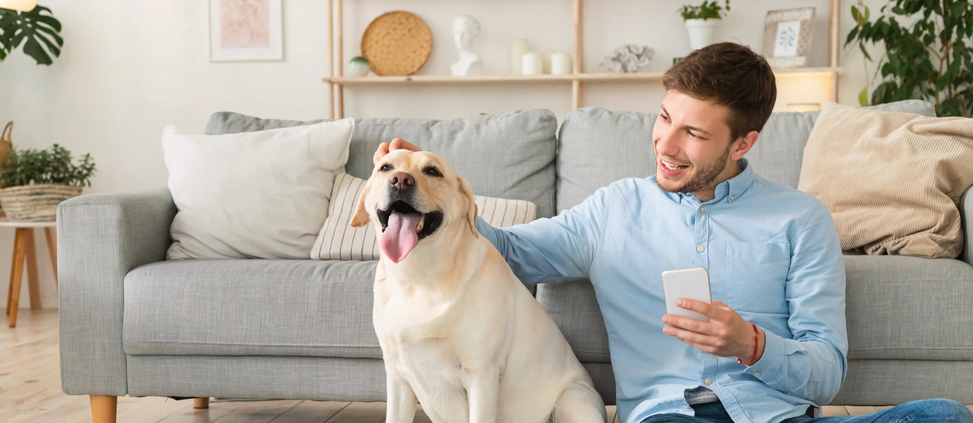 Mann sitzt mit Hund vor einem Sofa, hat sein Telefon in der Hand und lächelt.