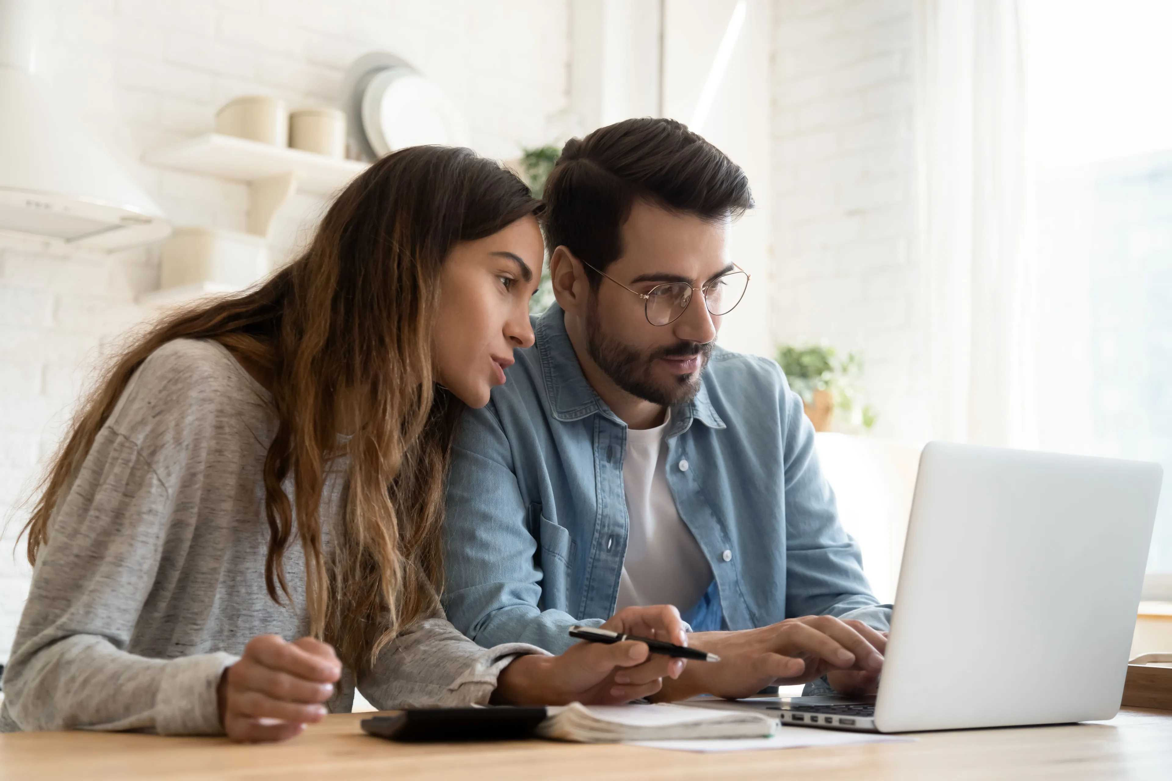 Ein Mann und eine Frau konzentrieren sich auf einen Laptop-Bildschirm und arbeiten an einem Holztisch in einer hellen, modernen Küche zusammen.