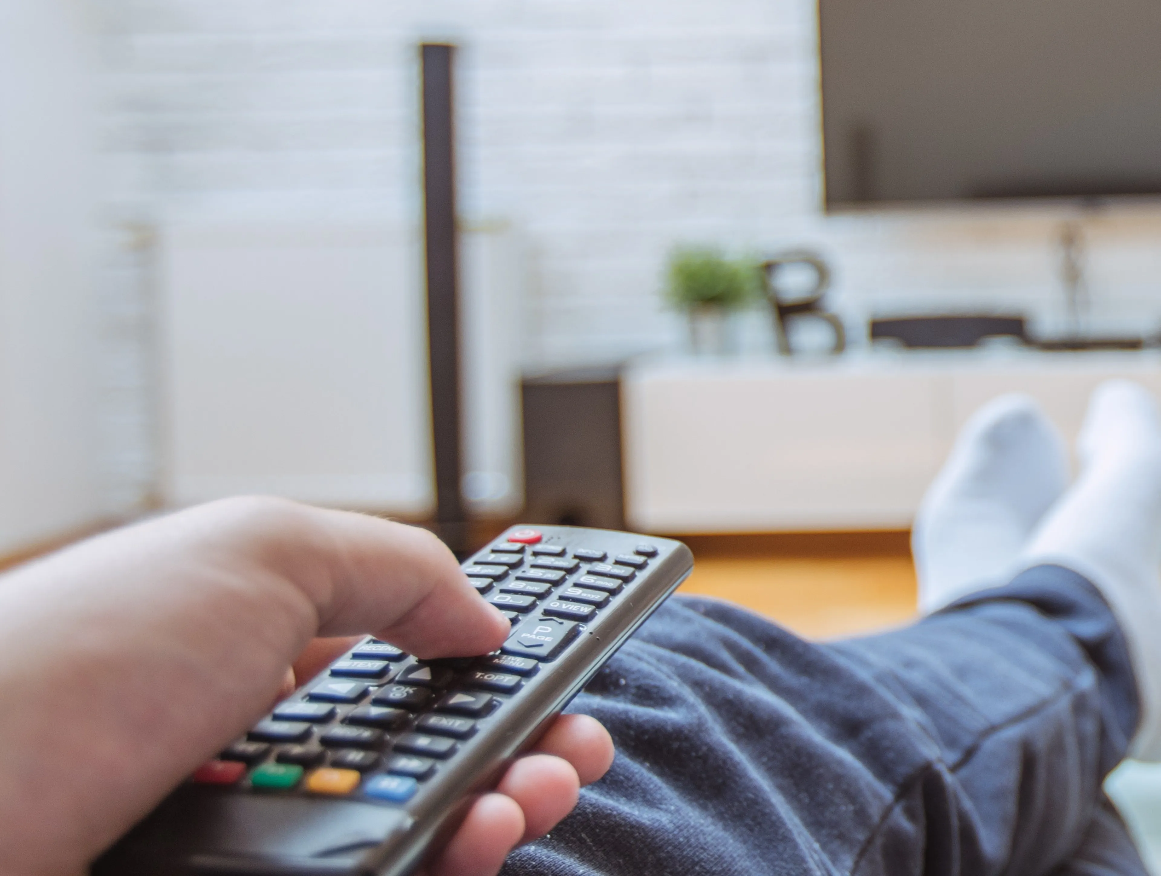 Person sitzt auf Sofa und hält die Fernbedingung richtung Fernseher