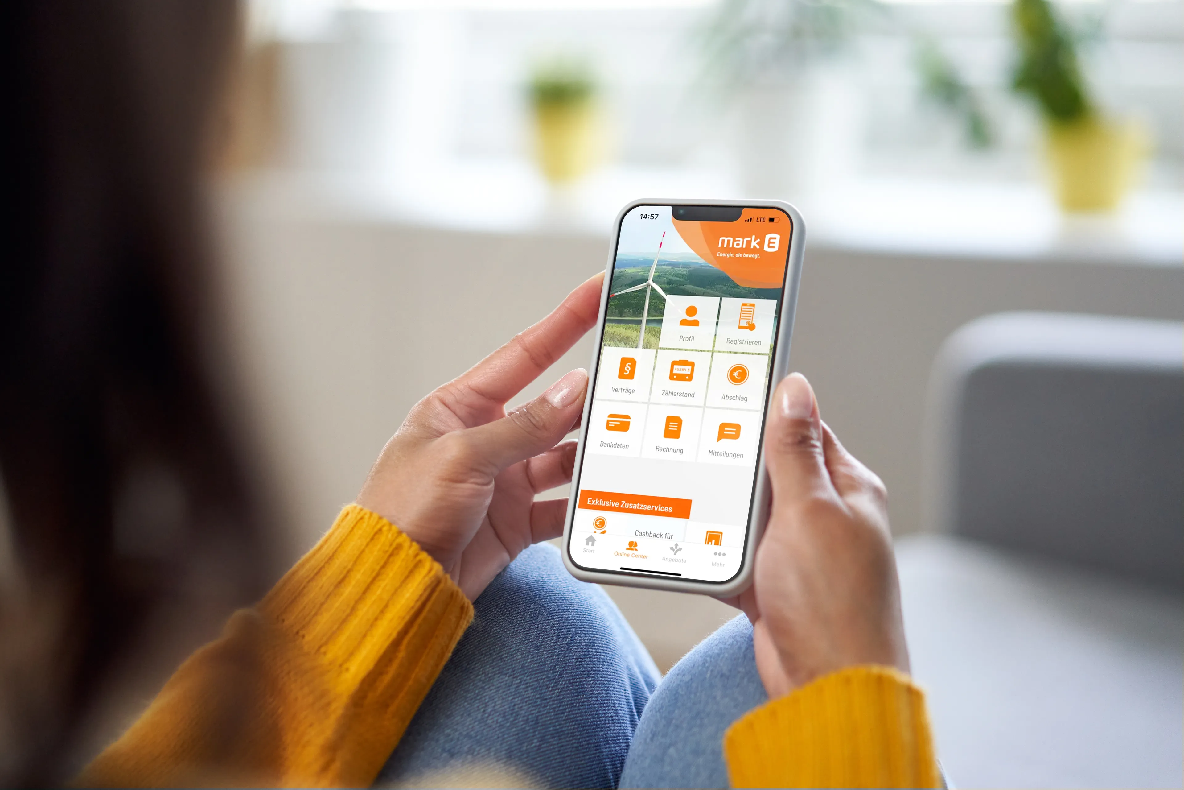 Person in gelbem Pullover, die ein Smartphone hält, auf dem eine App mit verschiedenen Symbolen angezeigt wird, sitzt bequem in einem Haus.