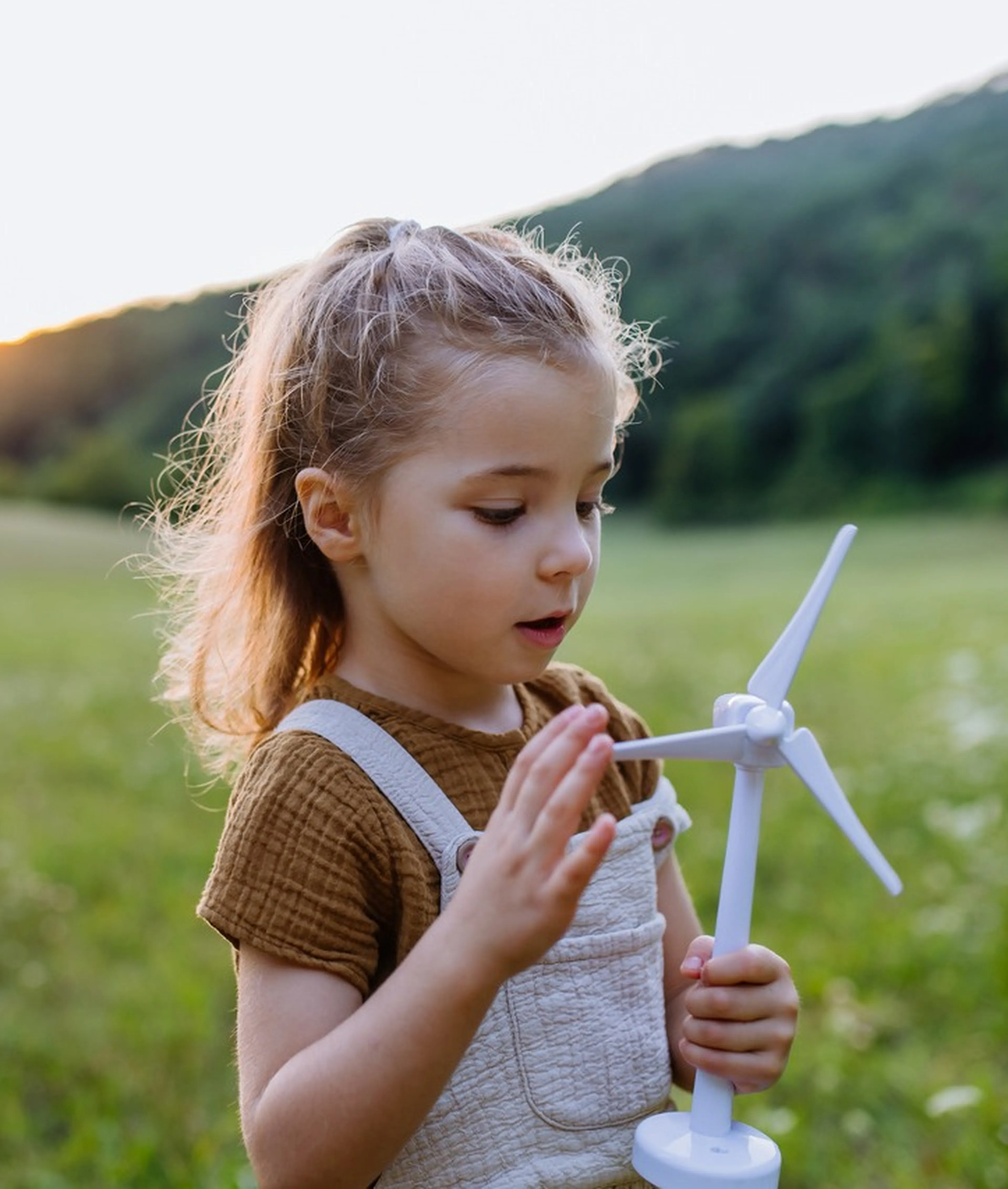 Ein junges Mädchen in einem braunen Hemd und einer Latzhose hält ein kleines Spielzeug-Windrad in einem grasbewachsenen Feld mit Hügeln im Hintergrund.