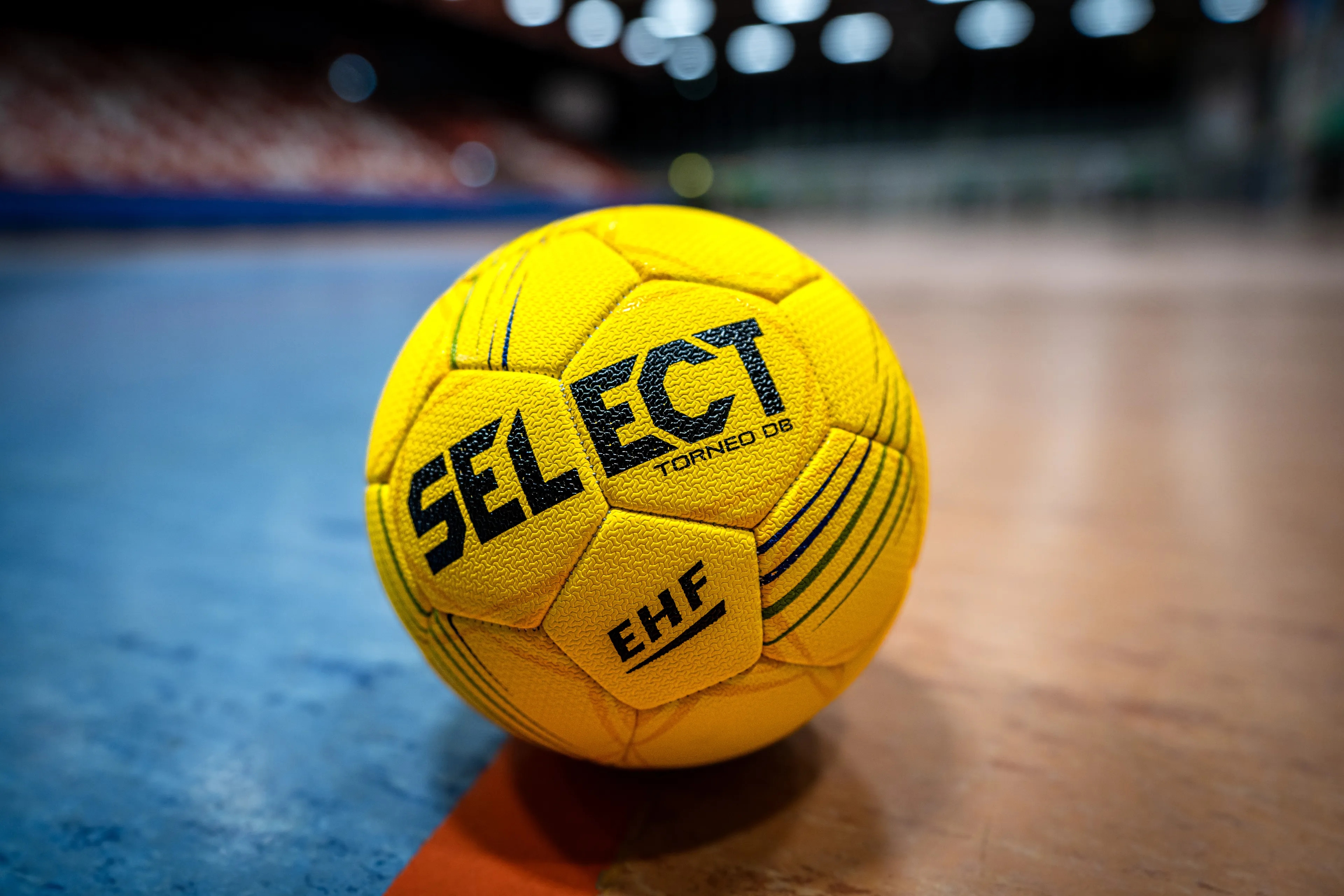 Ein gelber Handball mit „SELECT“- und „EHF“-Logo liegt auf einem Hallenplatz, im Hintergrund sind Sitzplätze und Lichter zu erkennen.