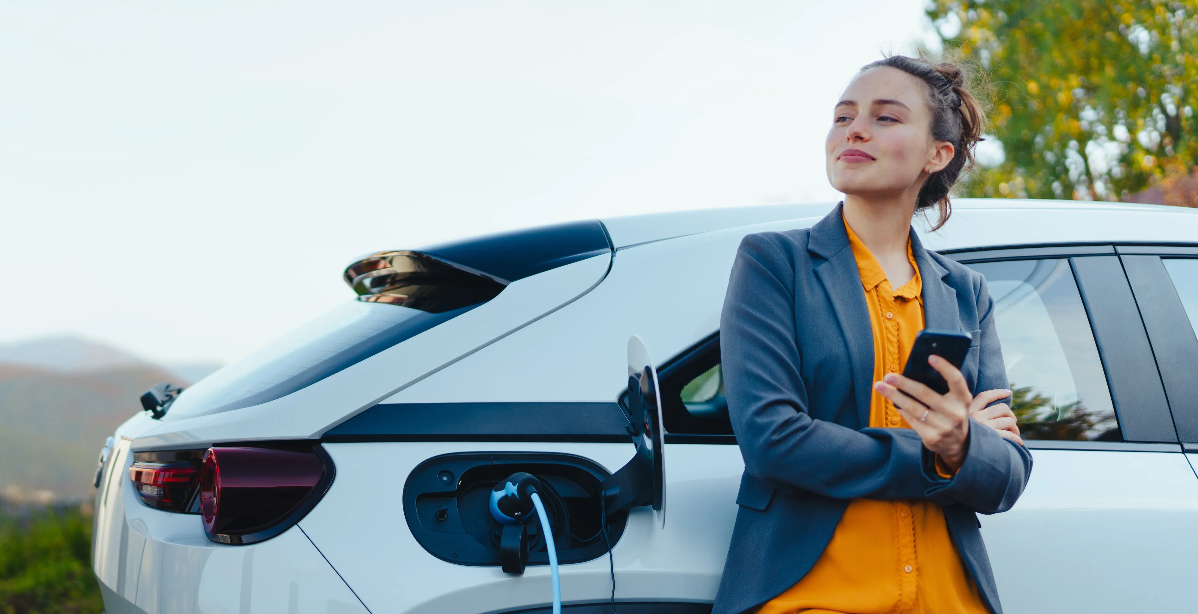 Lächelnde Frau lehnt an ladendem E-Auto mit Smartphone in der Hand