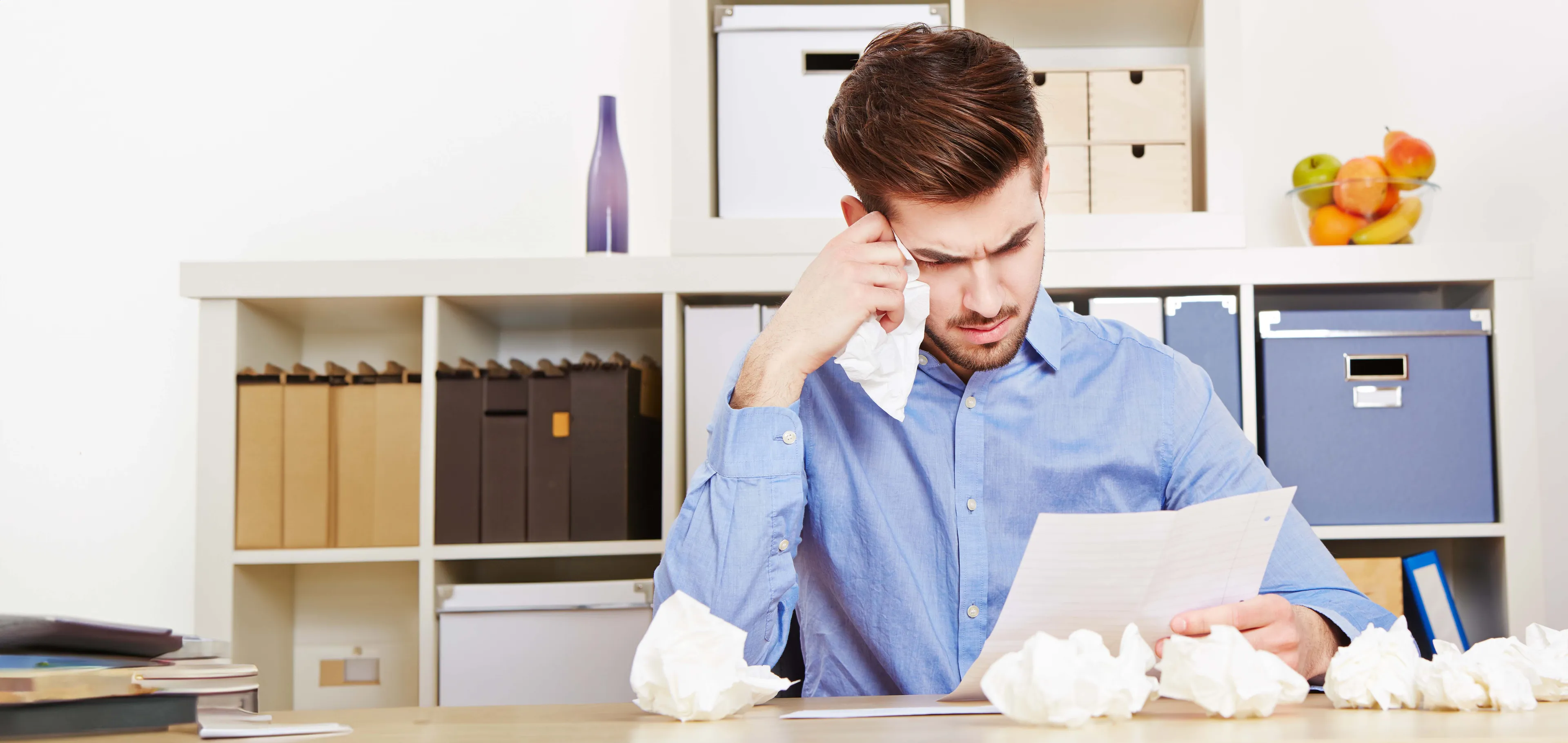 Mann in blauem Hemd hält Taschentuch und liest ein Dokument am Schreibtisch, umgeben von zerknitterten Papieren, mit Regalen und Aufbewahrungsboxen im Hintergrund.