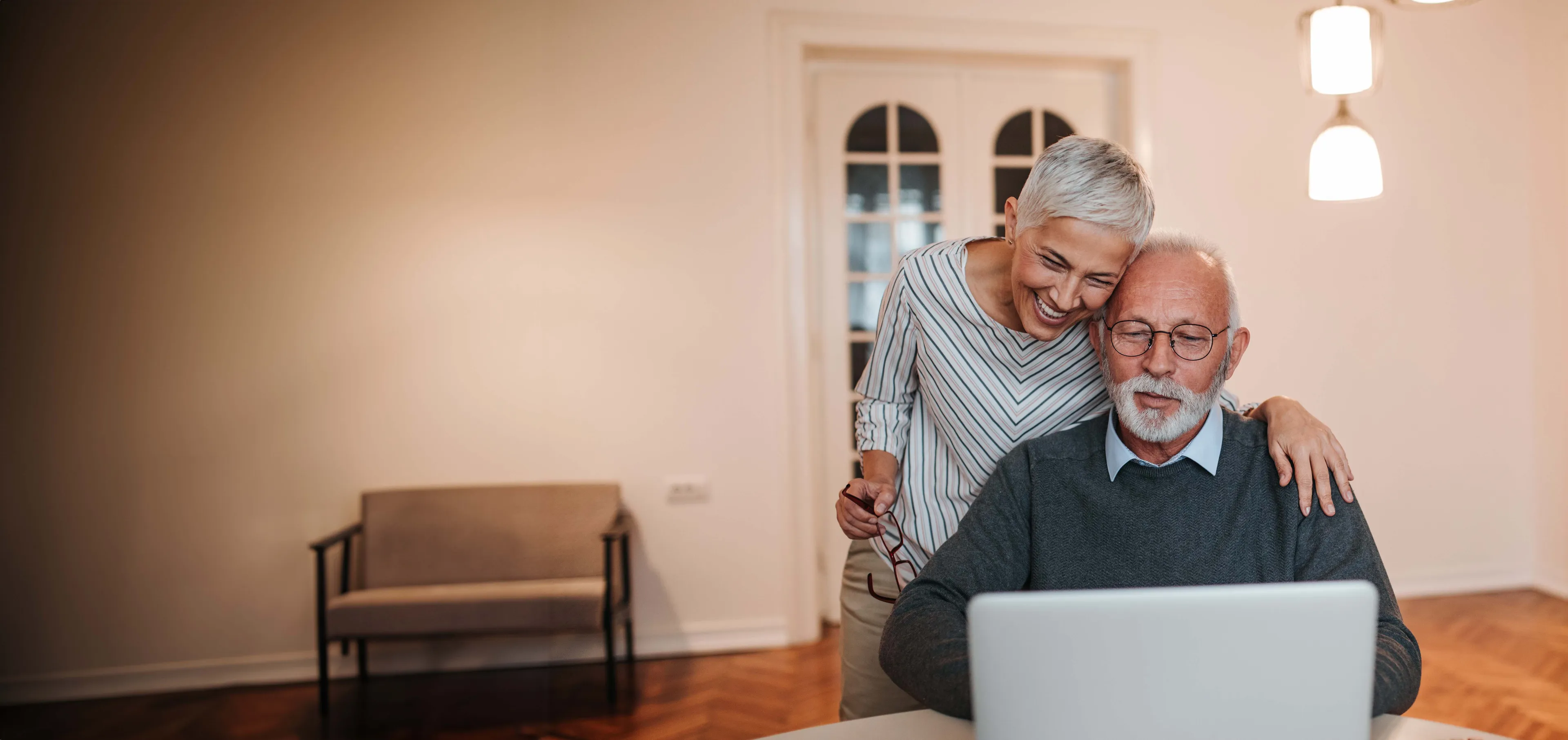 Älteres Paar lächelt, während es zu Hause einen Laptop benutzt; Frau steht und umarmt sitzenden Mann.
