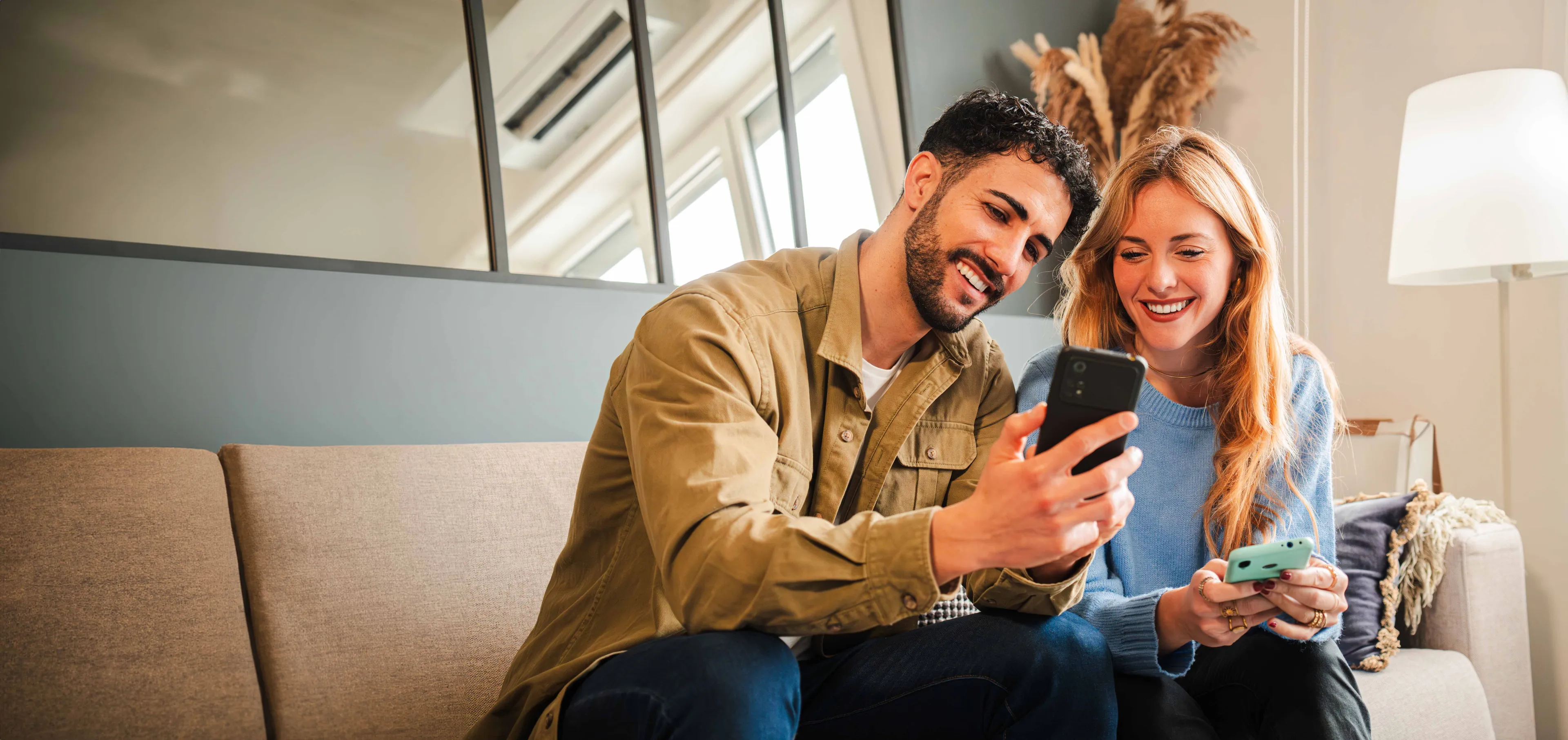 Zufriedenes Paar auf dem Sofa nutzt gemeinsam das Smartphone und lächelt dabei.